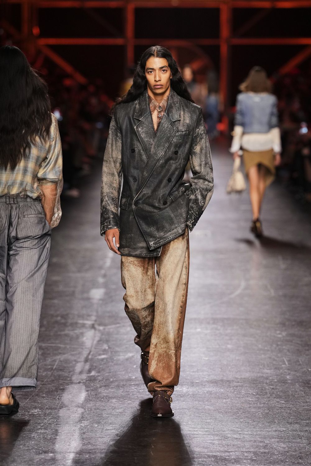 Dusty rose plaid shirt, top buttons open under the lapel. Black double-breasted blazer, heavily abraded to a chalky bloom. Wide cream trousers airbrushed brown at the hems and inner seams. Dark leather ankle boots with a brass ring at the vamp. For crossing the fairgrounds after rain, when the sawdust has turned to paste.