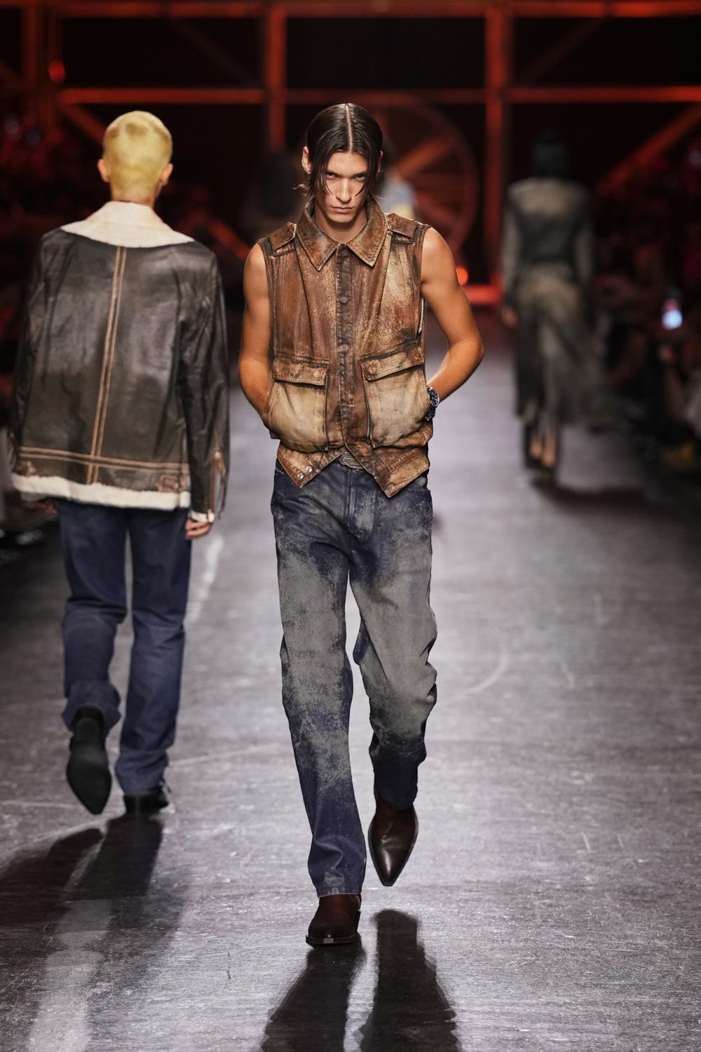 Burnished brown leather vest, mottled pale at the hem, flap pockets low. Indigo jeans dusted to ash through the thighs and knees. Dark square-toe boots peeking under the break. Crossing the fairgrounds after dark, generator hum and dirt still in the cuffs.