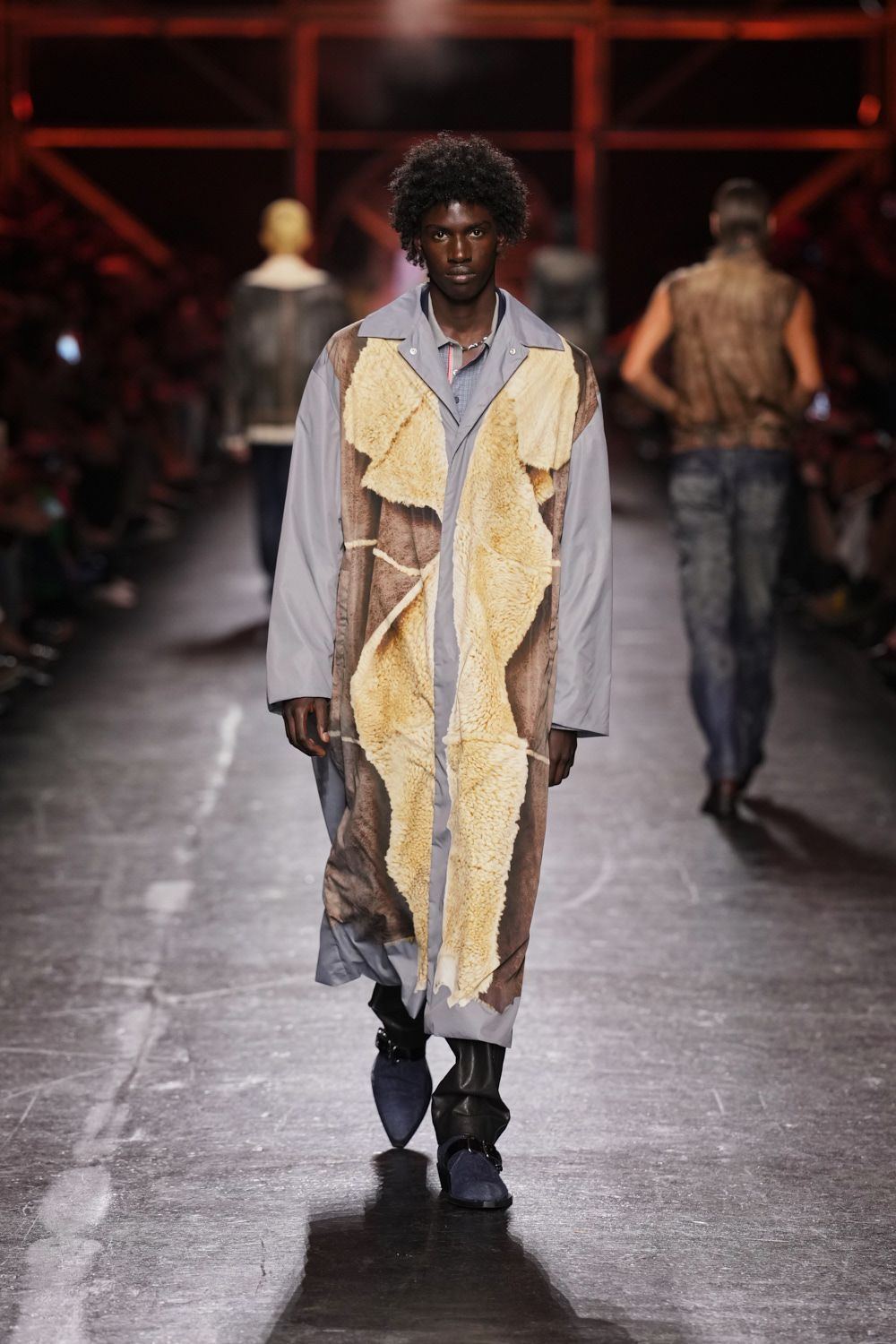 Dusty grey coat paneled with raw cream shearling hides, edges left jagged. Pale blue shirt collar spread over the coat neckline. Short silver chain at the throat. Black boots with navy suede spats buckled across the instep. For checking the stock fence at first light after a week of sleet.