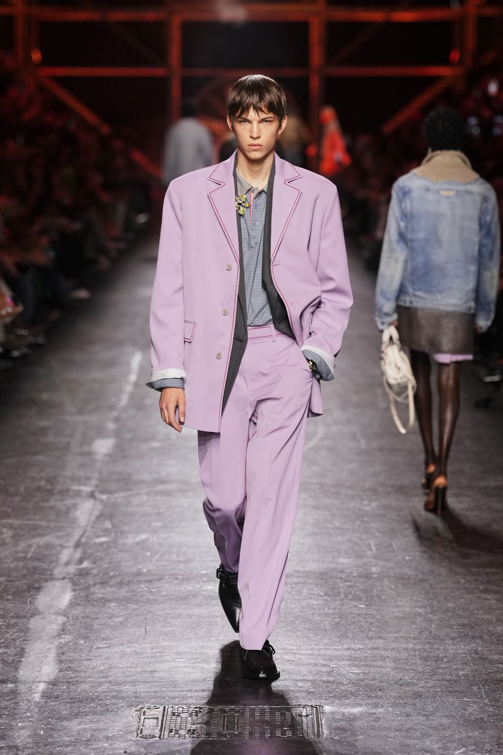 Oversized lilac blazer with burgundy blanket-stitch tracing the lapels. Grey microcheck polo, tipped placket pinned with a tiny corsage. Matching lilac trousers falling long over the shoe. Black square-toe derbies with a blunt shine. After the wedding lunch, cutting through the station before the flowers wilt.