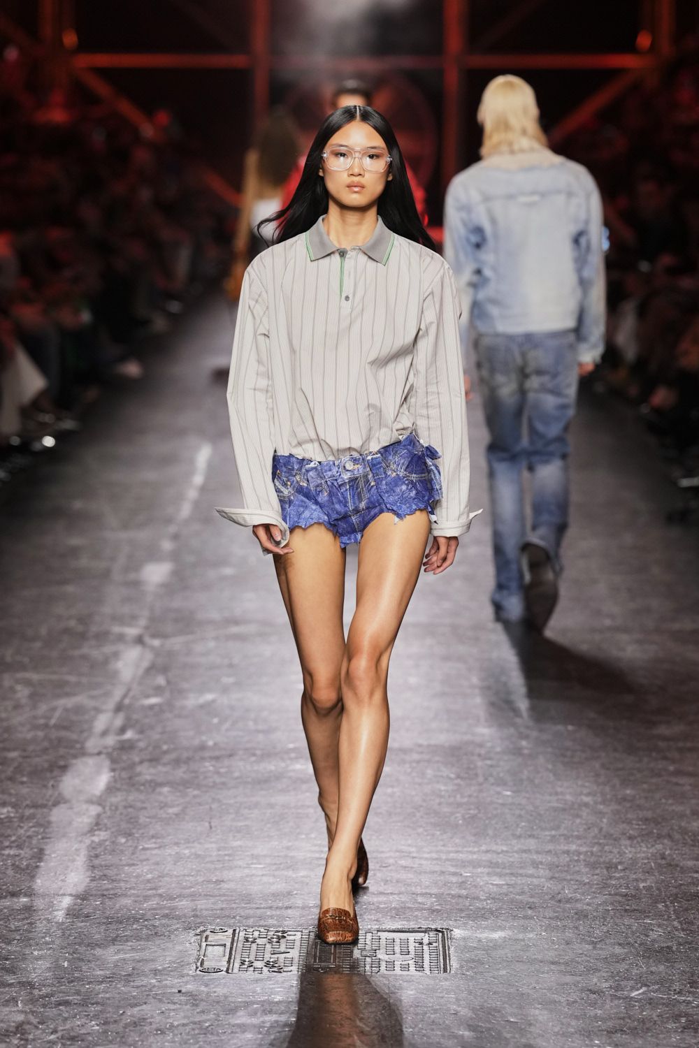 Clear aviator glasses with chunky transparent rims. Pale pinstriped polo, mint piping tracing the placket and collar. Ultra-low indigo denim shorts, shredded into a jagged handkerchief hem. Brown crocodile-embossed mules. Crossing a sticky arcade floor after the air-conditioning cut out.