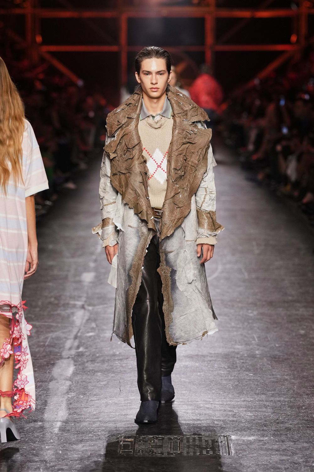Long coat of crumpled grey layers with ragged brown hide draped wide. Pale grey collar peeking cleanly above the knit. Oatmeal knit vest with a red zigzag argyle crossing the chest. Black leather trousers, one knee softly creased. Slate suede ankle boots. For the cold walk back from a fairground after rain.
