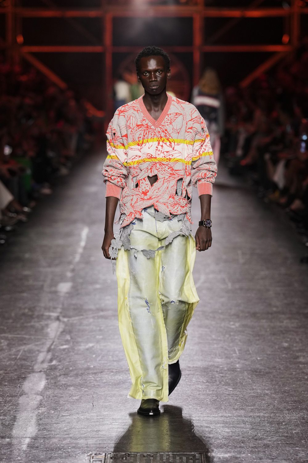 Grey V-neck knit with coral lily lines and a hacked-open waist. Pale yellow washed trousers veiled by a sheer trailing overskirt. Grey fabric flowers strung low across the hips. Square black boots. Whatever you wear home after sleeping through the station before dawn.