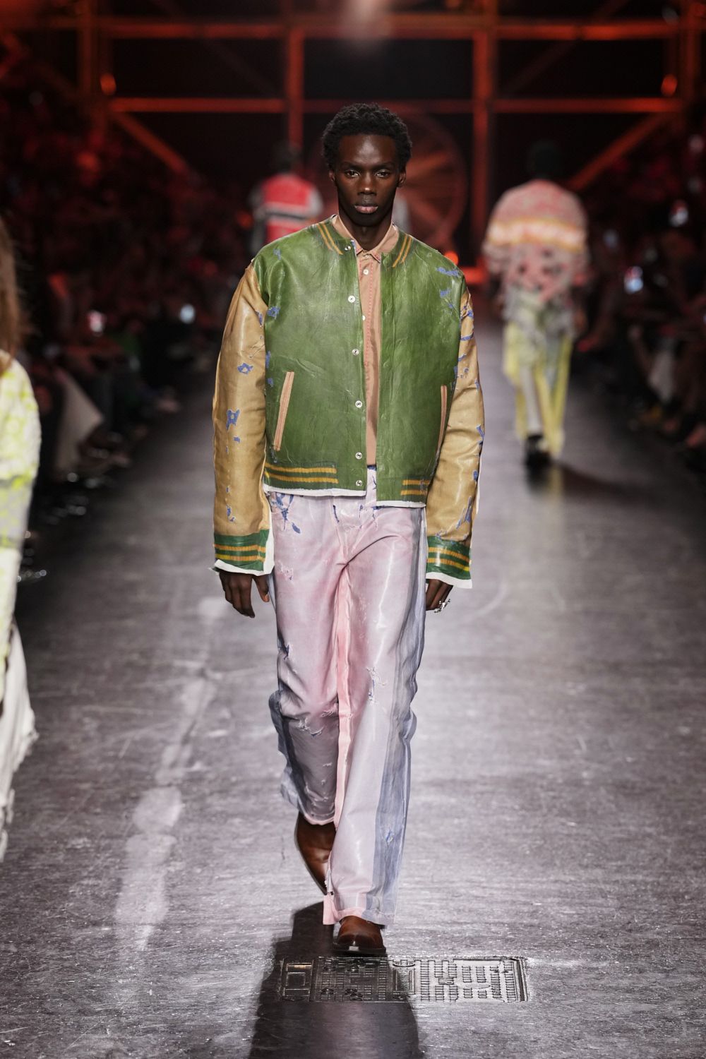 Bottle-green body with buttered leather sleeves, blue paint flecks and striped ribbing. Peach button-front shirt, collar points tucked under the jacket neck. Washed pink satin trousers, torn through in small scraped slashes. Brown square-toe shoes. Crossing the fairground after rain, tickets damp in a back pocket.