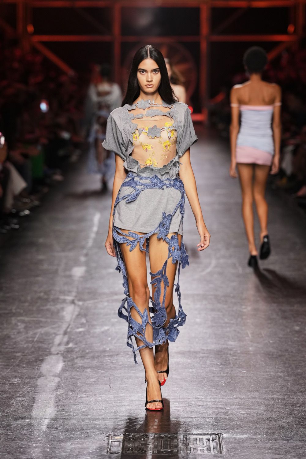 Grey jersey minidress cut away with appliqued floral straps and dangling blue cords. Nude mesh bodice speckled with yellow floral embroidery. Black thong sandals with a red-tipped pedicure. Crossing hot pavement after an art-school fundraiser let out.