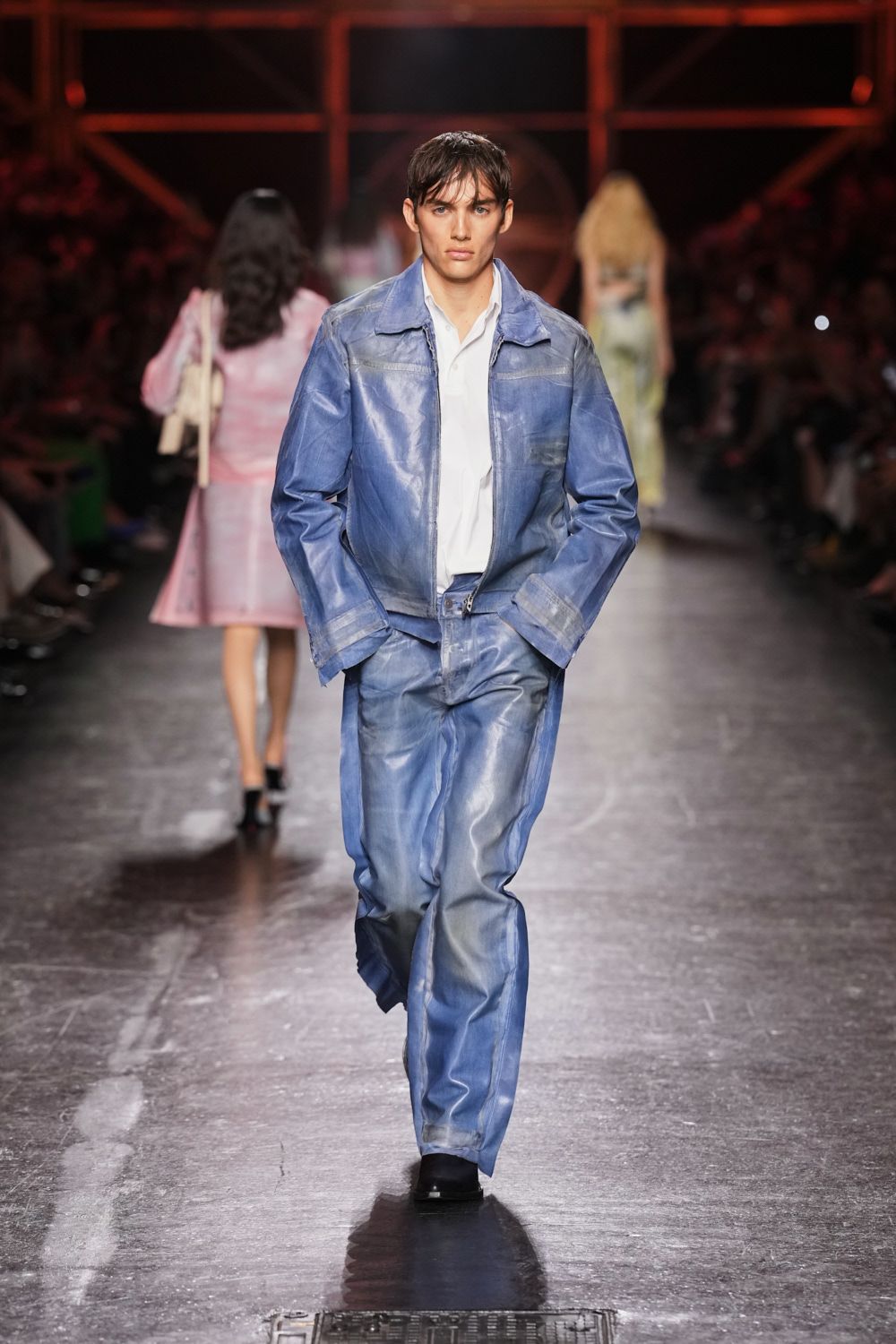 Washed blue leather jacket, seams ghosted pale and zip left slightly split. White shirt with a soft open collar and loose placket. Matching blue leather trousers, creased glossy through the thigh. Black square-toe shoes. Walking back from the marina after rain, salt drying on the railing.
