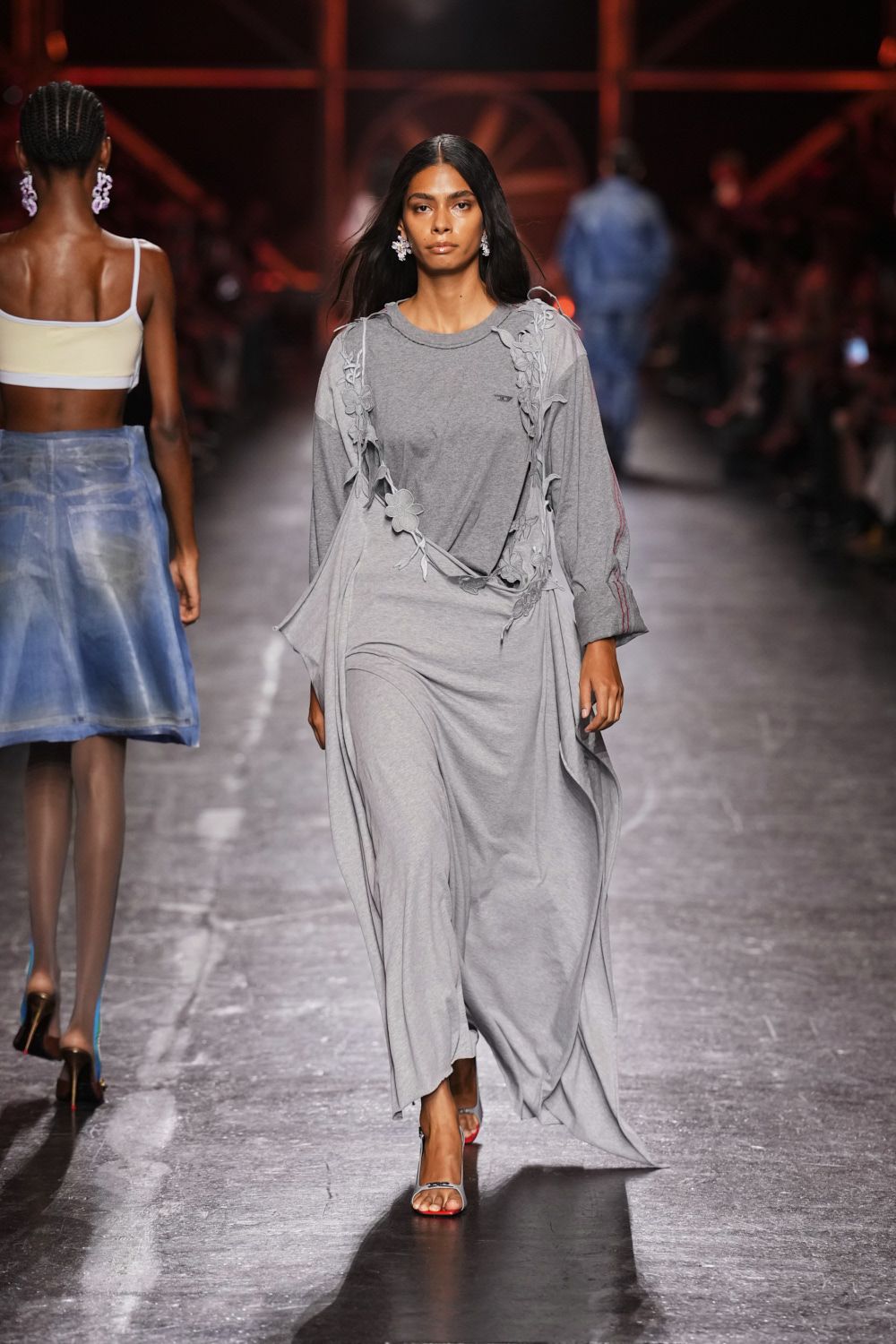 Heather-grey jersey maxi with a tee neckline and long trailing panels. Raw-edged floral cutouts climbing the shoulders and cinching one hip. Barely there heels with a scarlet sole flash at the step. Icy clustered drop earrings. Crossing a hotel lobby at dawn, still holding the night together.