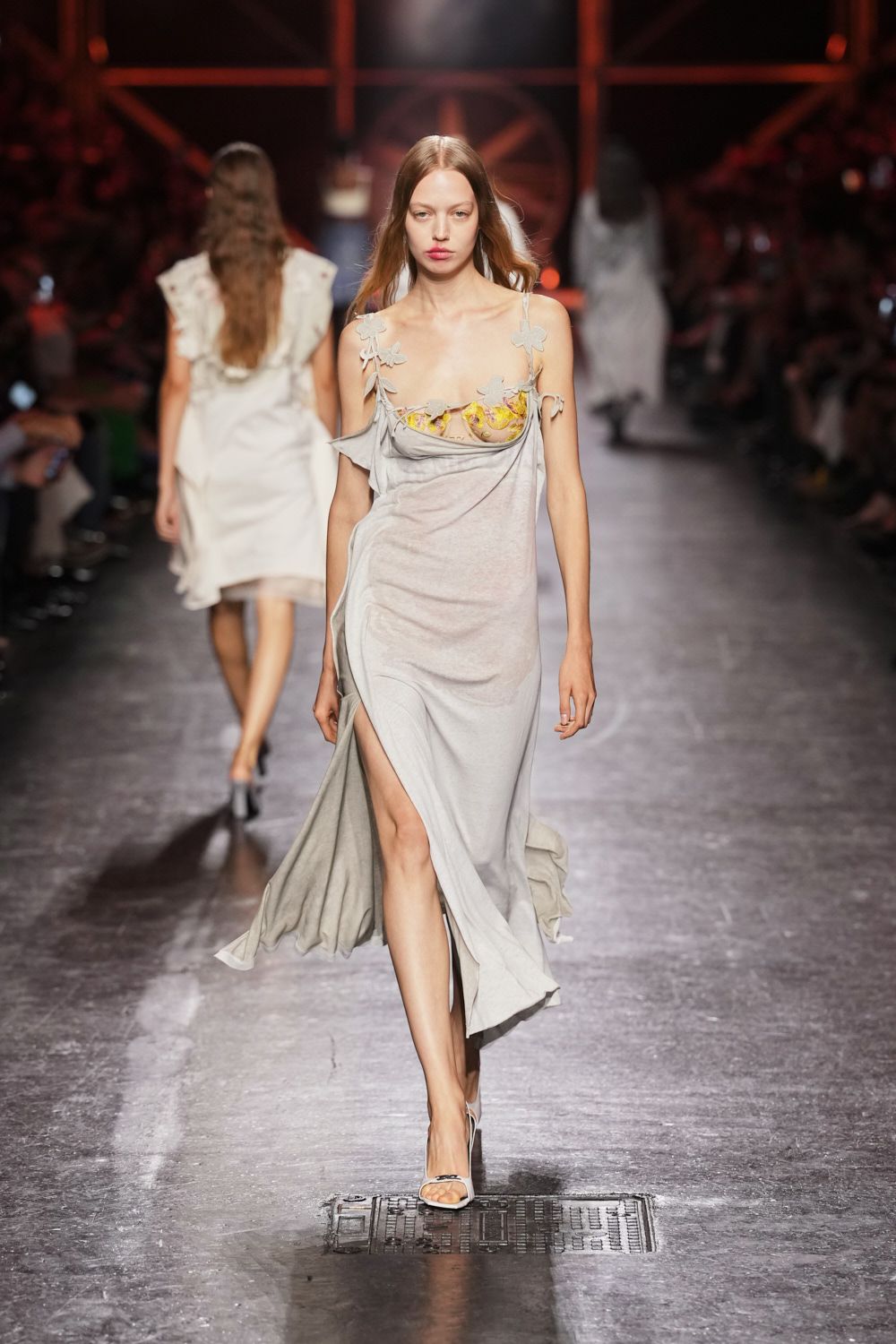 Ash-grey jersey slip dress draped off the shoulders, thigh slit swinging open. Buttercup embroidered cups exposed above the cowl, held by pale floral appliques. White ankle-strap sandals. Crossing a hotel ballroom after the wedding cake has been cut.