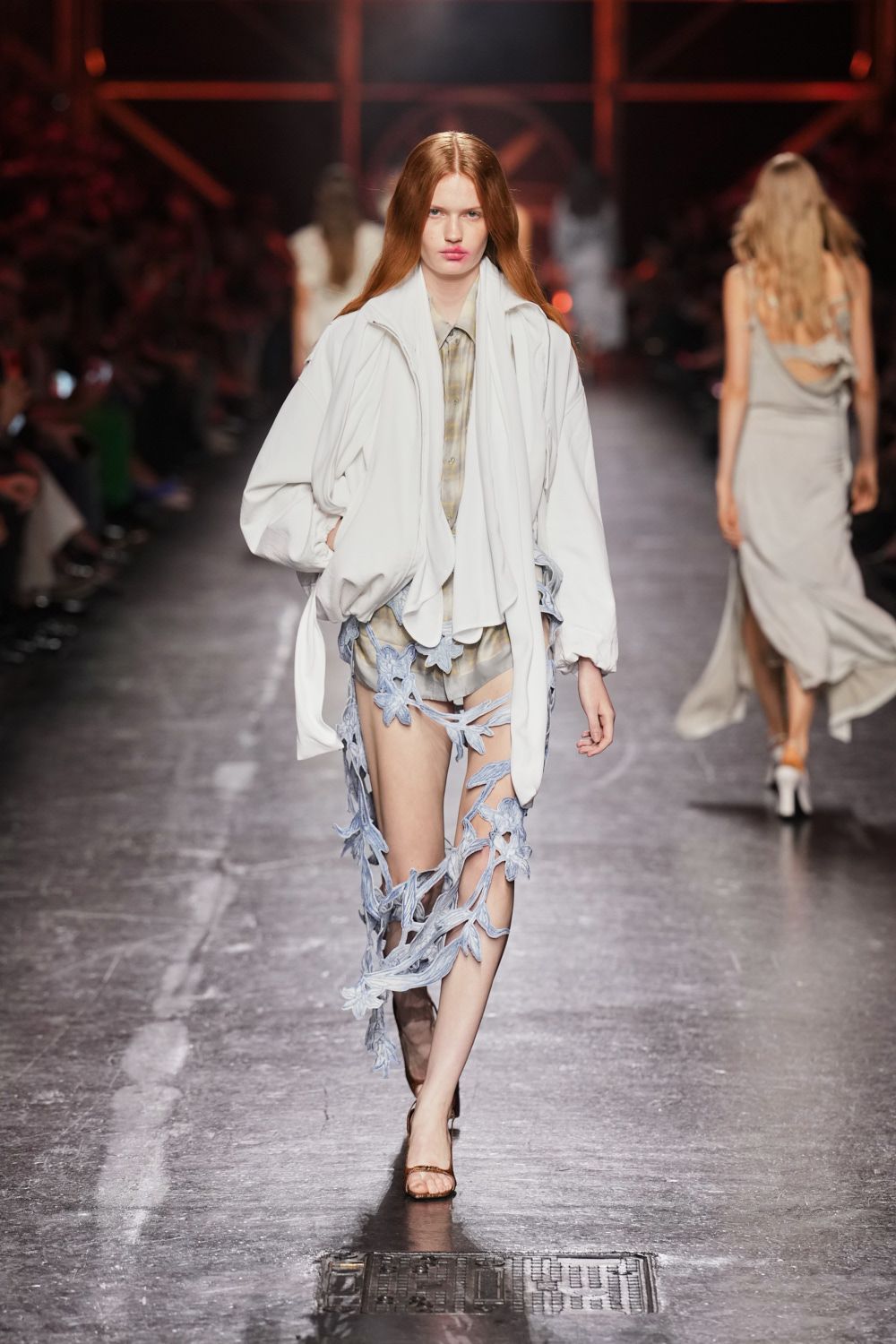 Cloud-grey blouson with a soft funnel neck and ballooned cuffs. Washed plaid shirt peeking out beneath the zip front. Pale satin shorts, loose through the thigh. Powder-blue floral strands climbing one leg like torn lace. Brown thong sandals. Crossing wet concrete after dawn with petals stuck to your calves.