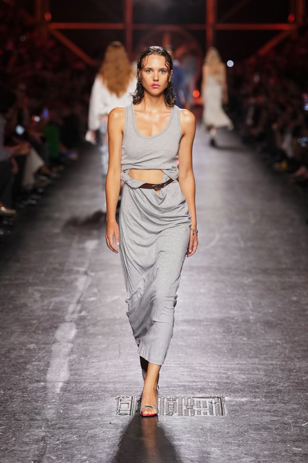 Heather-grey jersey tank dress twisted open at the waist. Thin brown leather belt buckled through the cutout. Clear-strap heeled sandals with a coral-tinted sole edge. Walking back from the beach with salt still drying at your ribs.