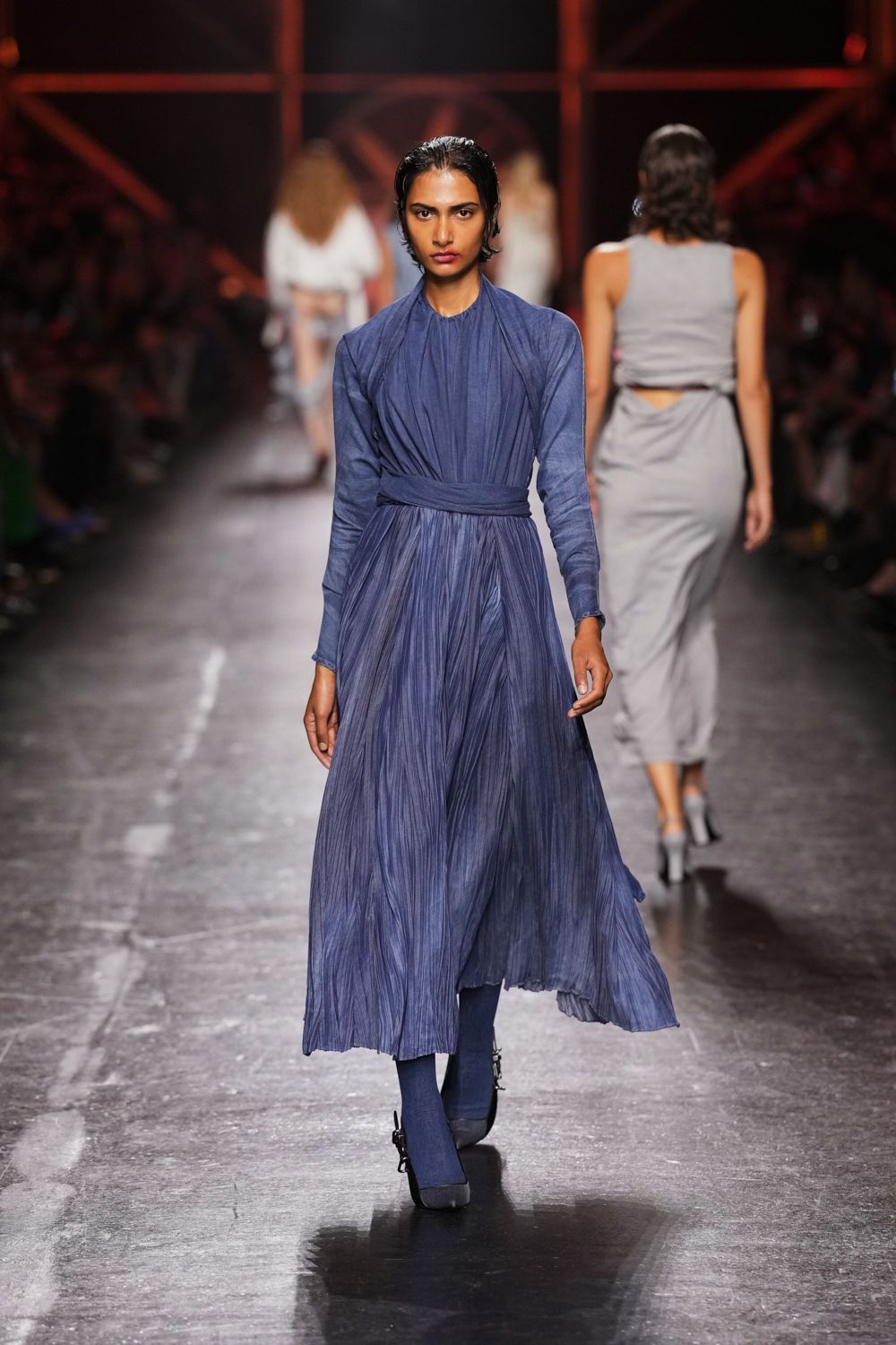 Dusty indigo dress in crushed pleats, neckline softly gathered to the throat. Self-fabric sash wrapped flat and knotted low at the waist. Matching opaque indigo tights. Black platforms with a squared toe and silver side buckles. Crossing a museum atrium just before the rain blows in.
