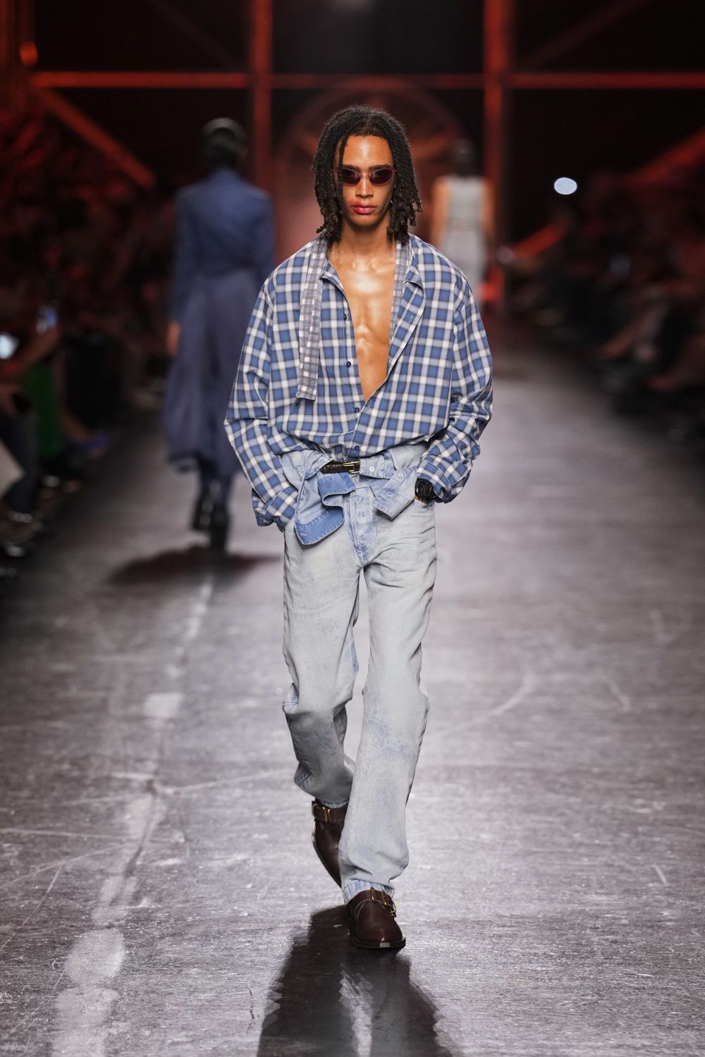 Narrow plum-tinted lenses in a thin oval frame. Oversized blue plaid shirt worn open, a scarf panel hanging from the collar. Bleached straight-leg jeans, waistband cinched with a slouched denim tie. Dark leather mules with a gold horsebit across the vamp. Walking out of a beach motel at noon in yesterday's clothes.