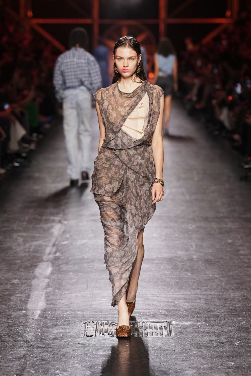 Smoke-brown floral chiffon dress, draped in overlapping panels with a cream ruffle inset. Dangling clustered earrings. Stacked jewel-tone bracelets. Brown embossed leather pumps. Walking back from a wedding through streets still warm after rain.