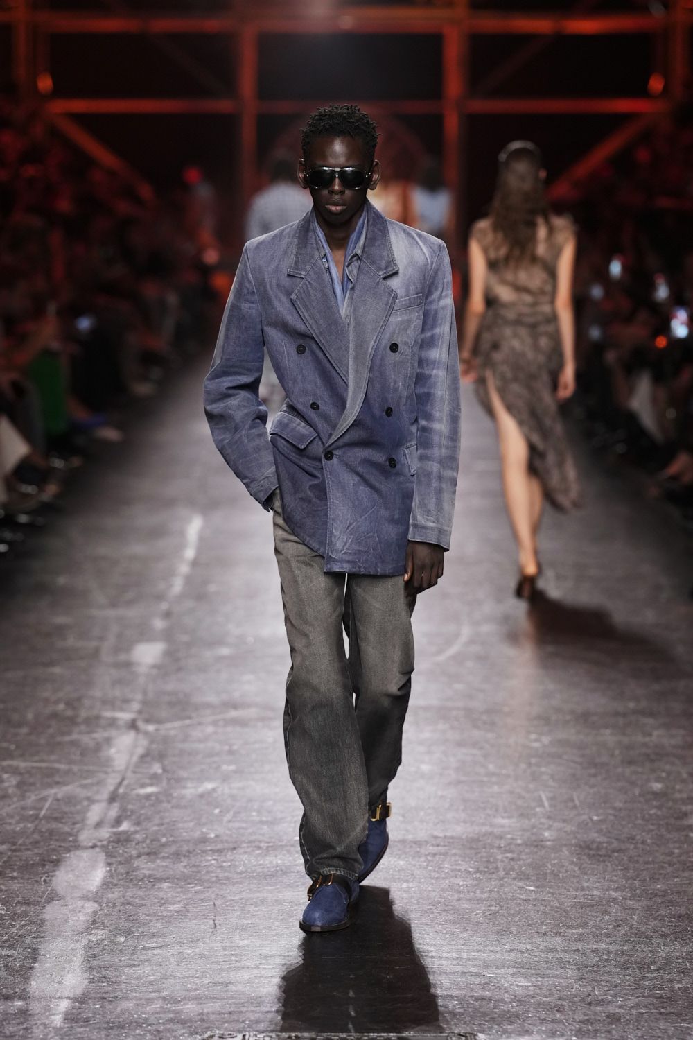 Oversized black shield sunglasses. Dusty periwinkle velvet double-breasted blazer, lapel edge curling open. Slate blue shirt left loose at the throat. Washed grey jeans falling straight with a soft break. Blue suede ankle boots with a square gold bit. Crossing a casino floor at dawn, jacket still holding last night's smoke.