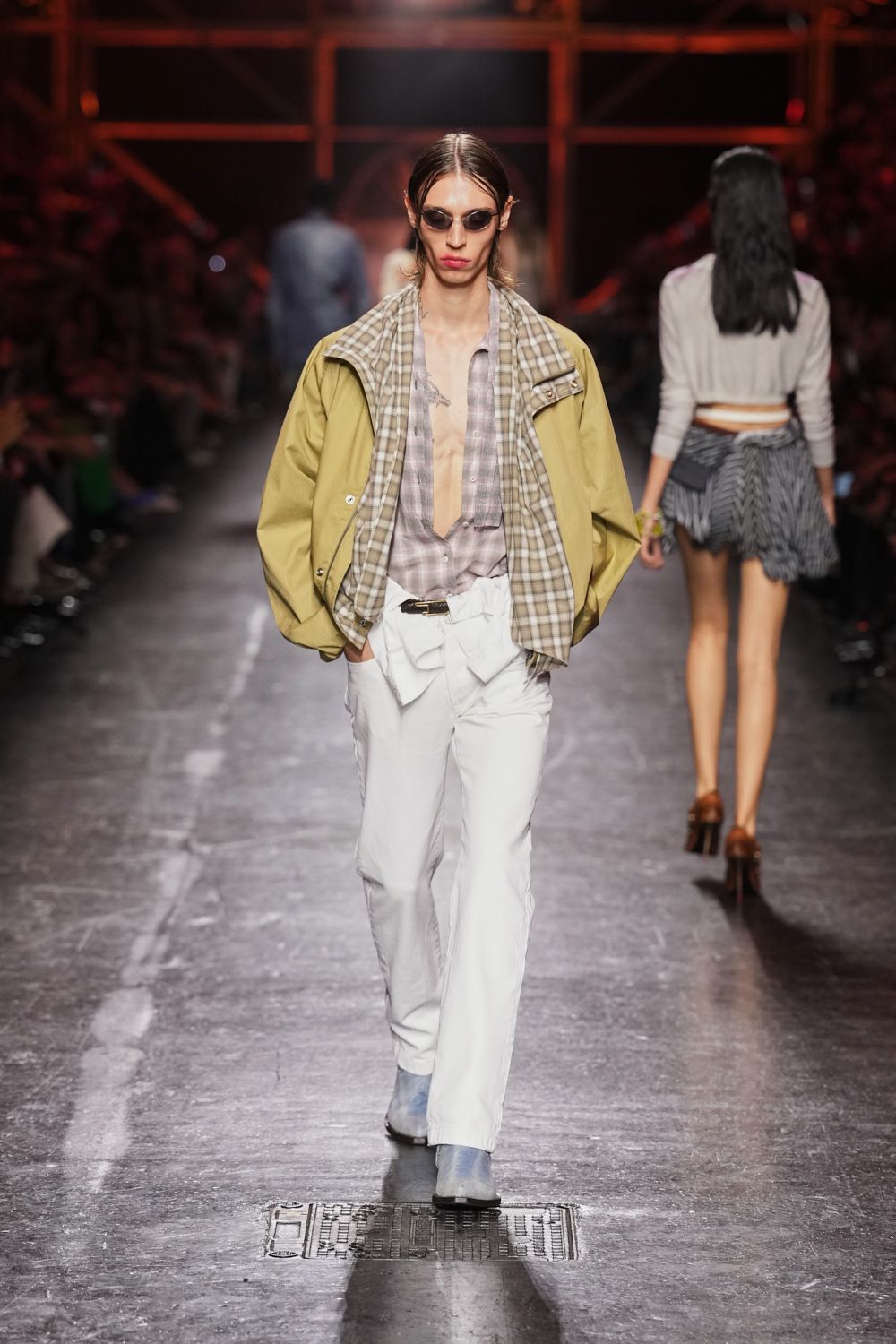 Small smoked oval lenses with thin metal rims. Light olive windbreaker, beige plaid lining turned outward at the collar. Pale mauve plaid shirt hanging open to a plunging V. White trousers with a paperbag waist and tied self belt. Powder blue suede ankle boots. Walking under station lights after missing the last coastal train.