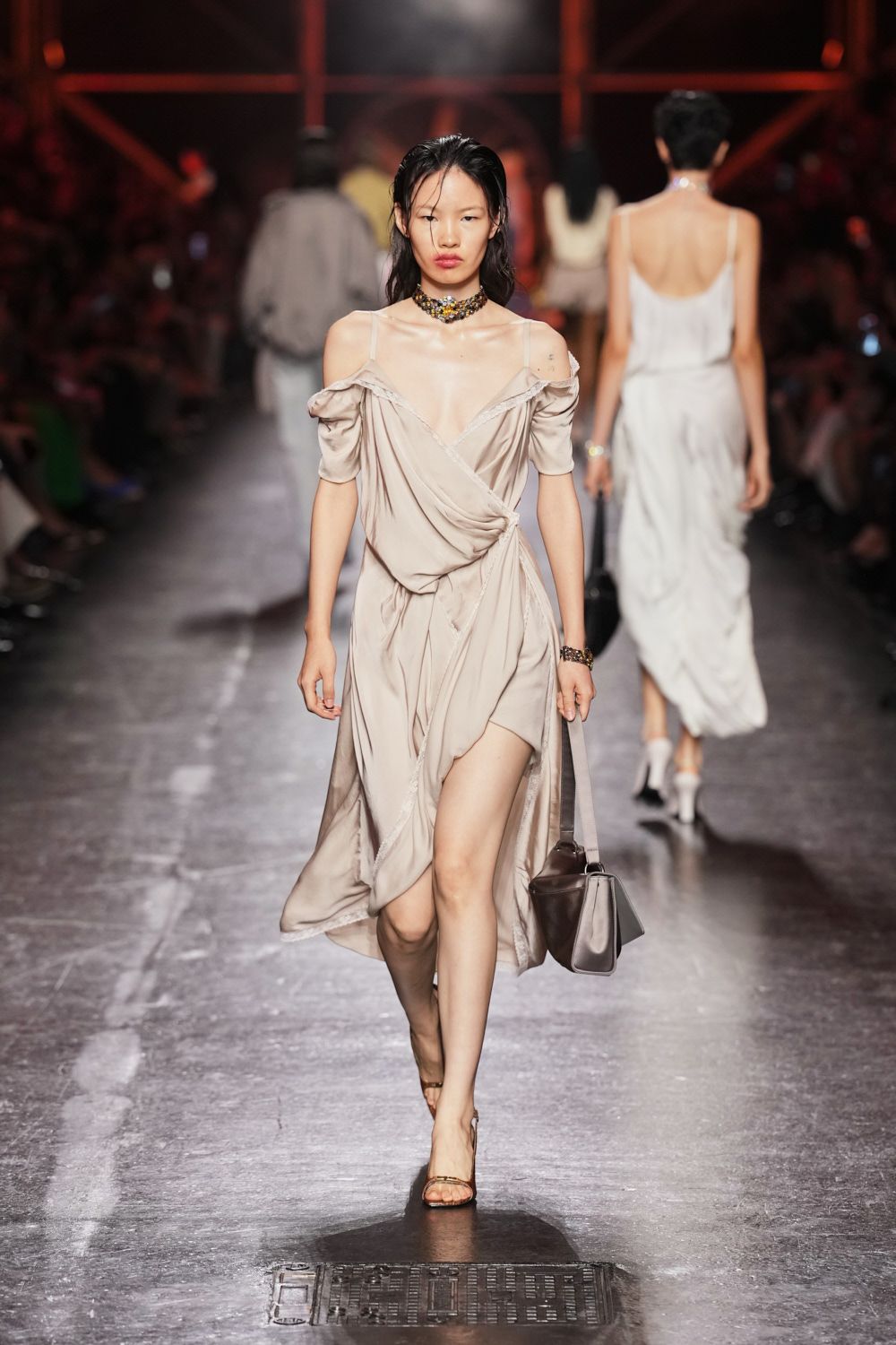 Dove satin wrap dress, slipped off the shoulders with raw-edged drape. Clustered jewel choker, dark stones packed tight at the throat. Matching jeweled cuff. Taupe leather shoulder bag with a rigid boxed side panel. Barely there nude sandals. Crossing a hotel lobby at dawn, carrying last night's shoes in your hand.