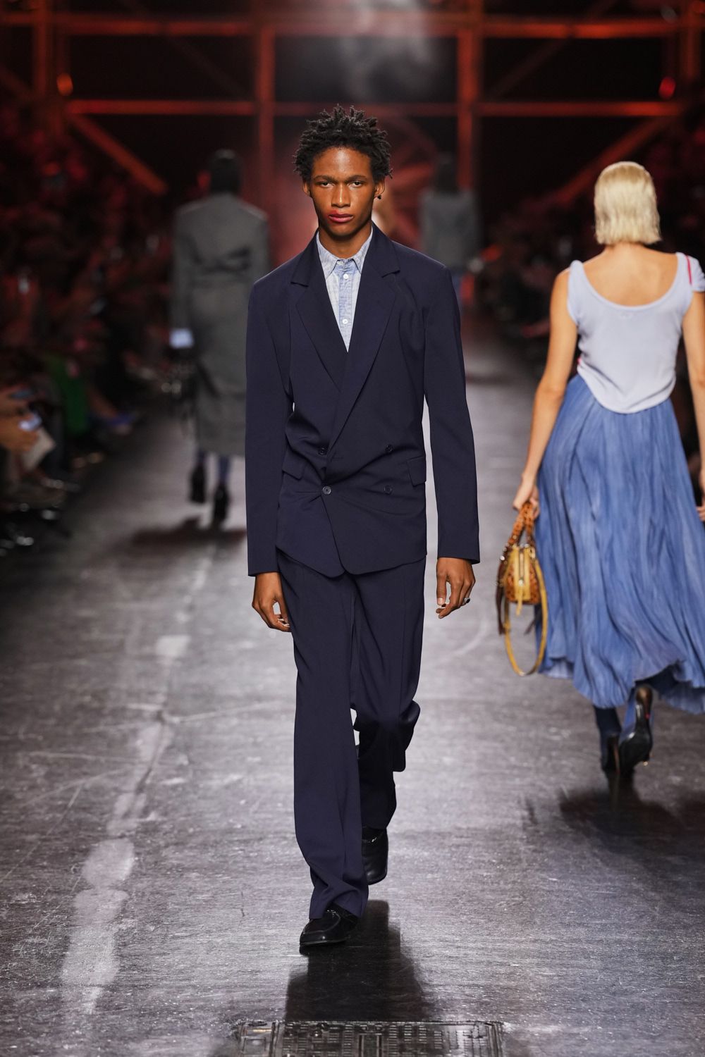Ink navy blazer with a skewed double-breasted button stance. Pale blue shirting, tiny white buttons fastened to the throat. Matching straight trousers breaking softly over the shoes. Black square-toe shoes with a low shine. First train after rain, carrying your suit through a station still smelling of metal.