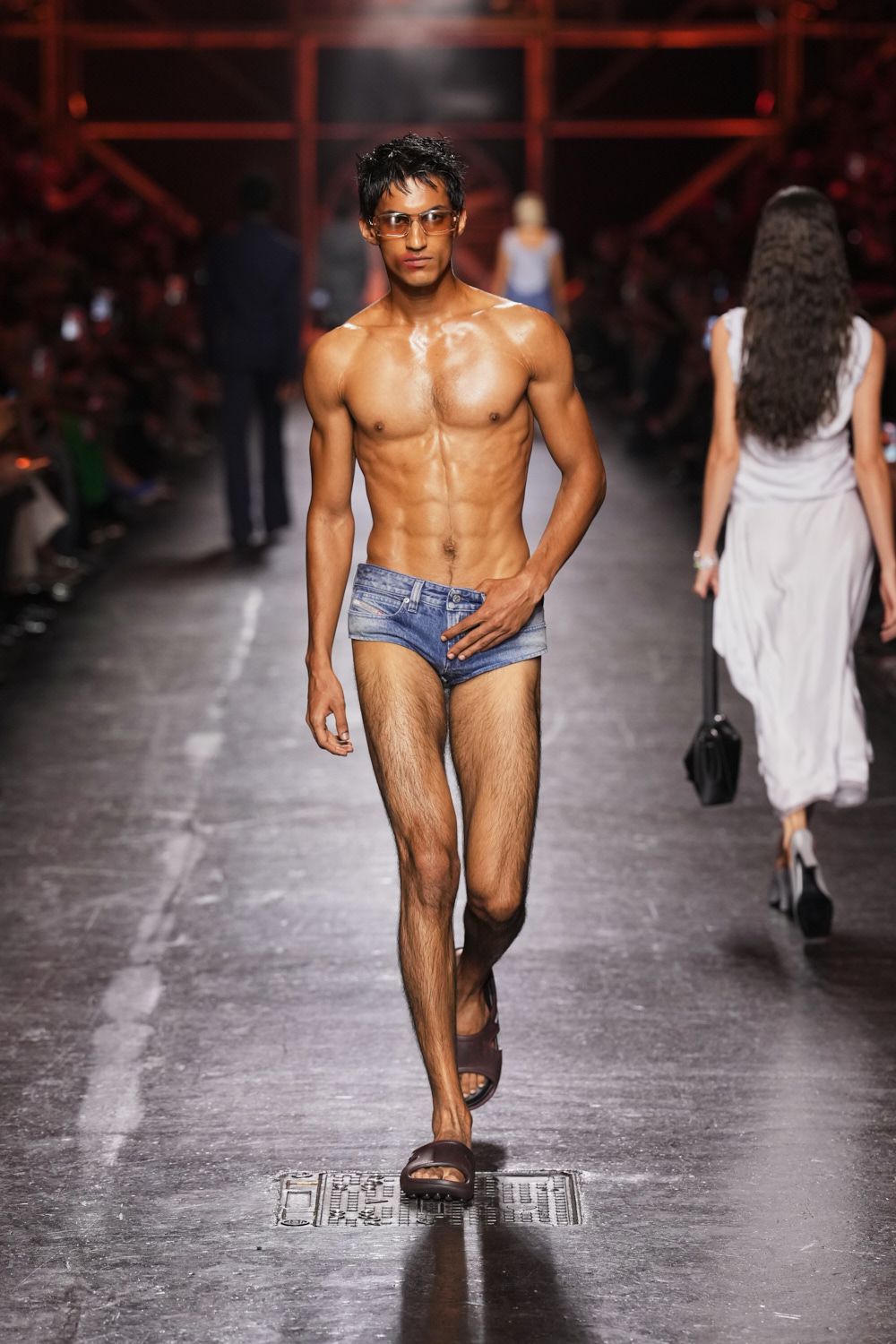 Amber wraparound lenses with a thin metallic bridge. Low-rise denim briefs, whiskered and faded, fly seam left visible. Brown molded slides with broad matte straps. Pool deck after midnight, skin still salted and not yet dry.