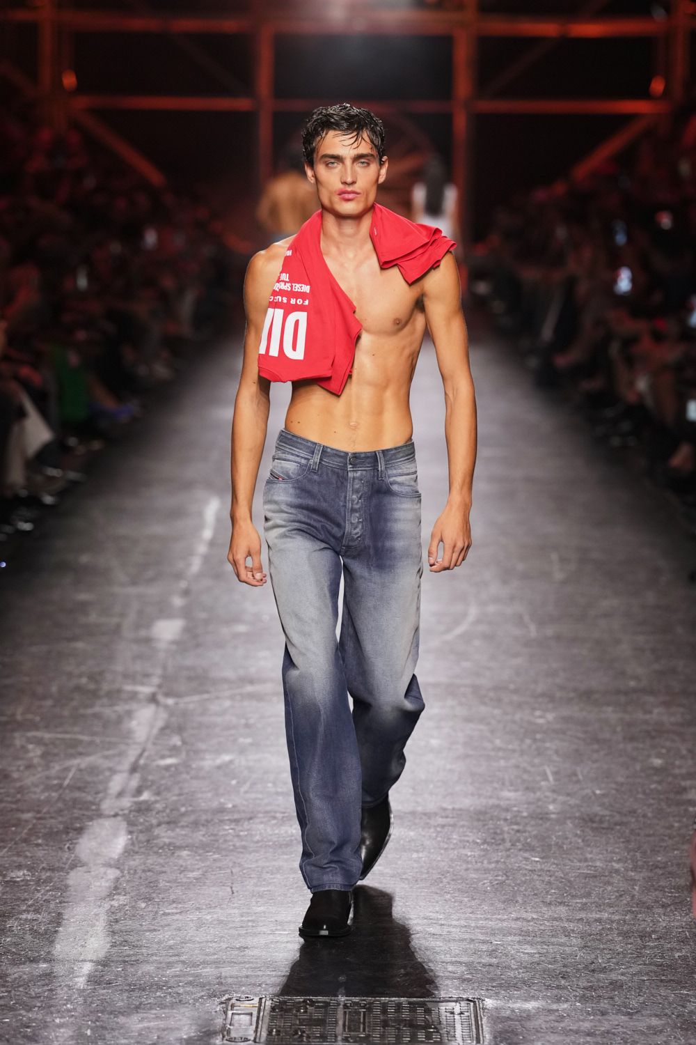 Red slogan tee slung off one shoulder, collar twisted into a loop. Faded slate denim jeans with whiskering and a low, loose rise. Black square-toe boots. Walking back from the river at dawn in whatever stayed dry.