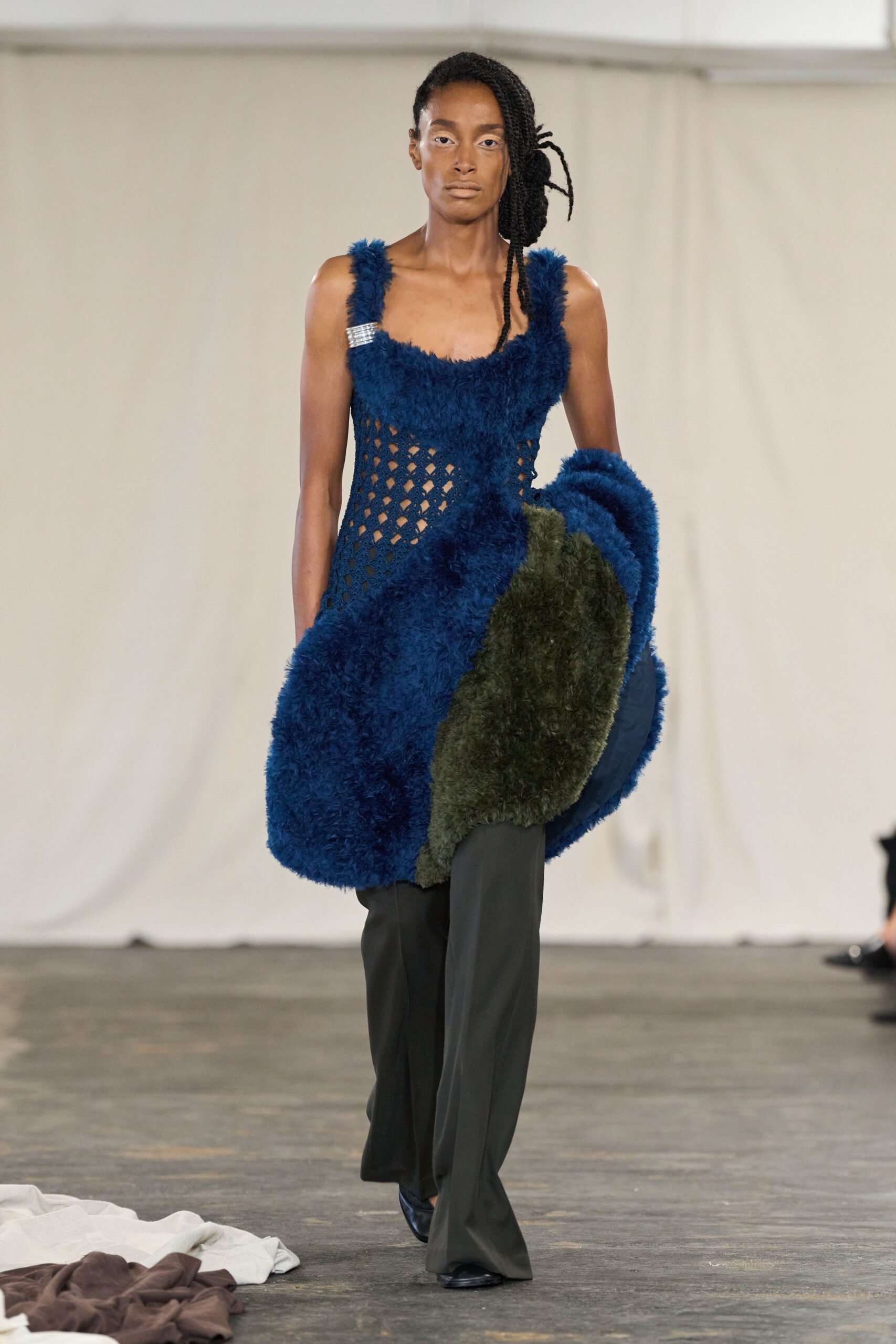 Deep blue shag-knit dress with open lattice sides and a rhinestone strap clip. Olive and cobalt faux-fur coat slung in the crook of one arm. Black fluid trousers breaking softly over the shoe. Black pointed shoes. Crossing a drafty art school hall with someone else's blanket over your arm.
