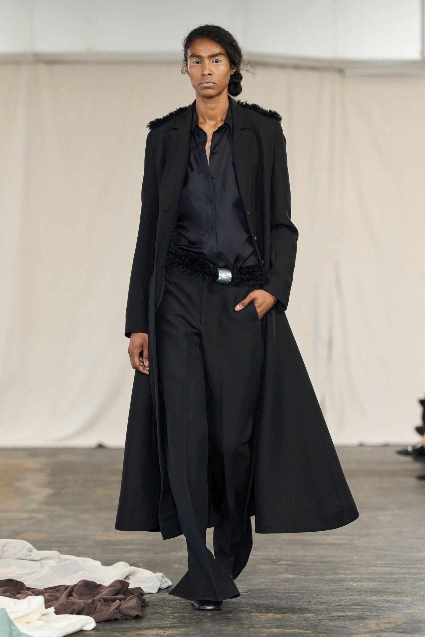 Long black coat with fuzzy tufts running over the shoulders. Black satin shirt, unbuttoned low with a soft collapsed placket. Black feathery belt closed by a dull silver oval plaque. Wide black trousers breaking heavily over the shoe. For crossing a hotel lobby before dawn with yesterday still on you.