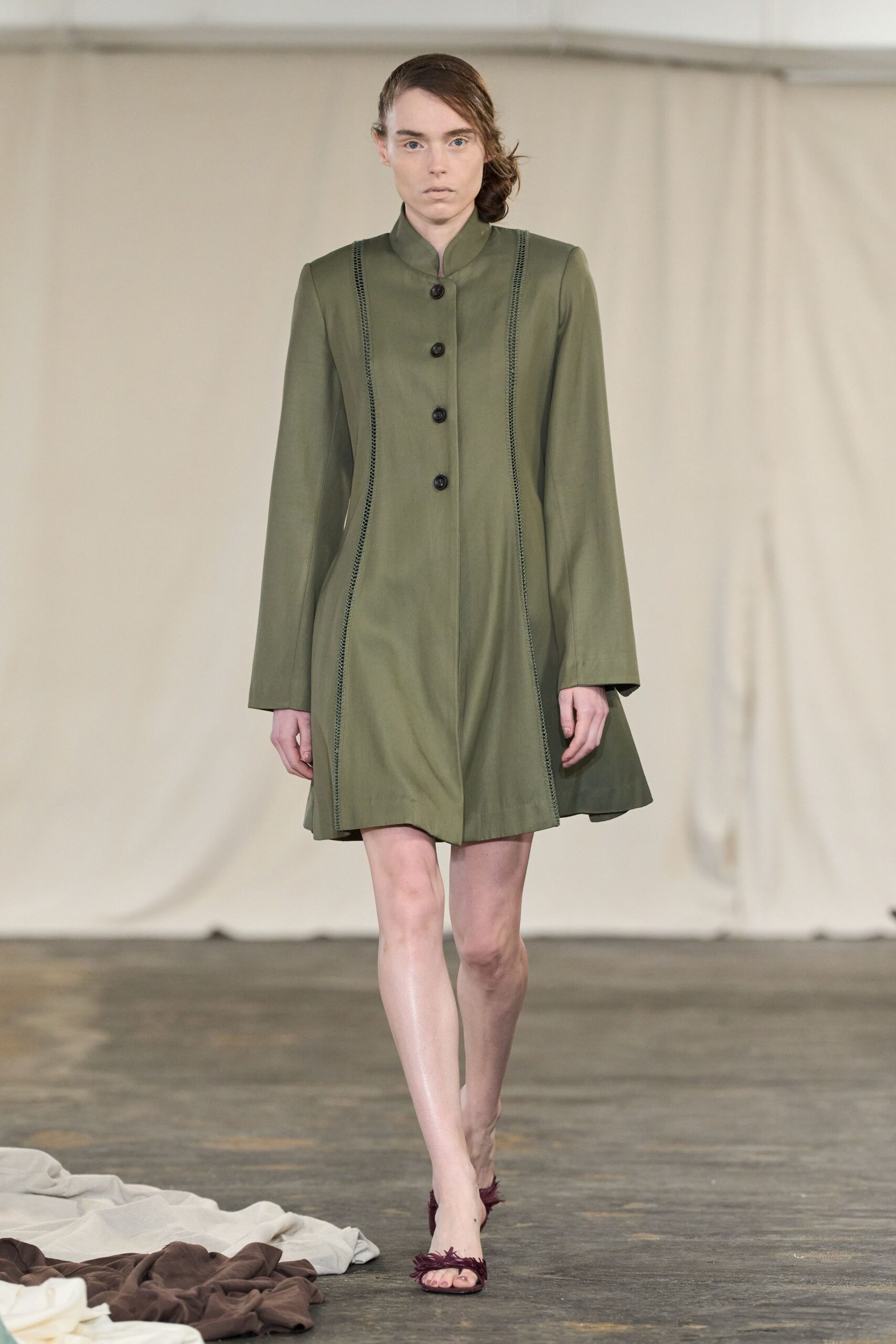Olive coatdress with a stand collar and laddered seams curving inward. Burgundy sandals topped with wiry feather fringe across the toes. Crossing a museum atrium before the rain starts again.