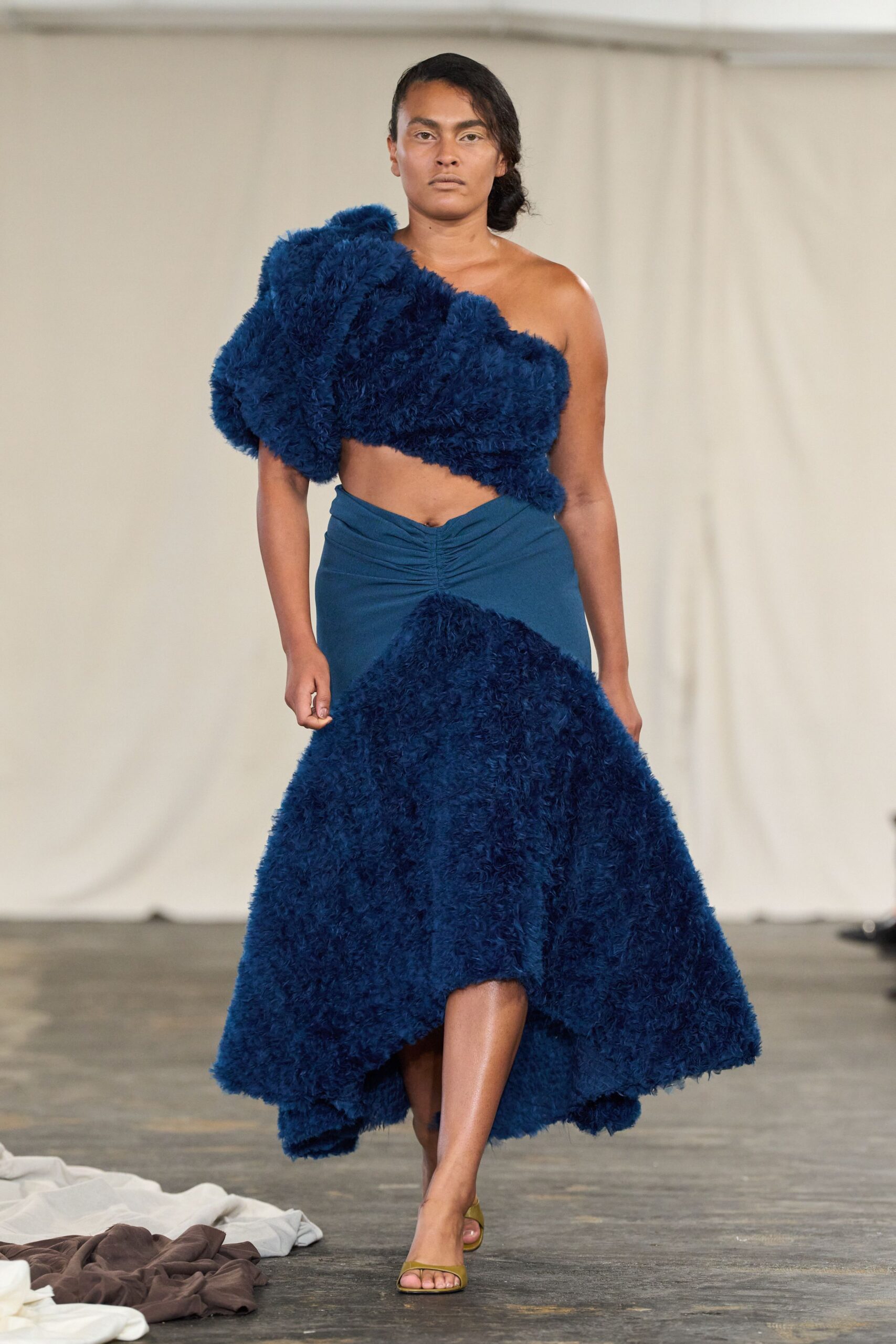 One-shoulder cropped top in dense cobalt fleece, swelling into clouded volume. Deep teal jersey skirt, ruched at the waist with a shaggy fleece godet. Olive satin slide sandals. Crossing a museum atrium after rain, carrying half the sky with you.