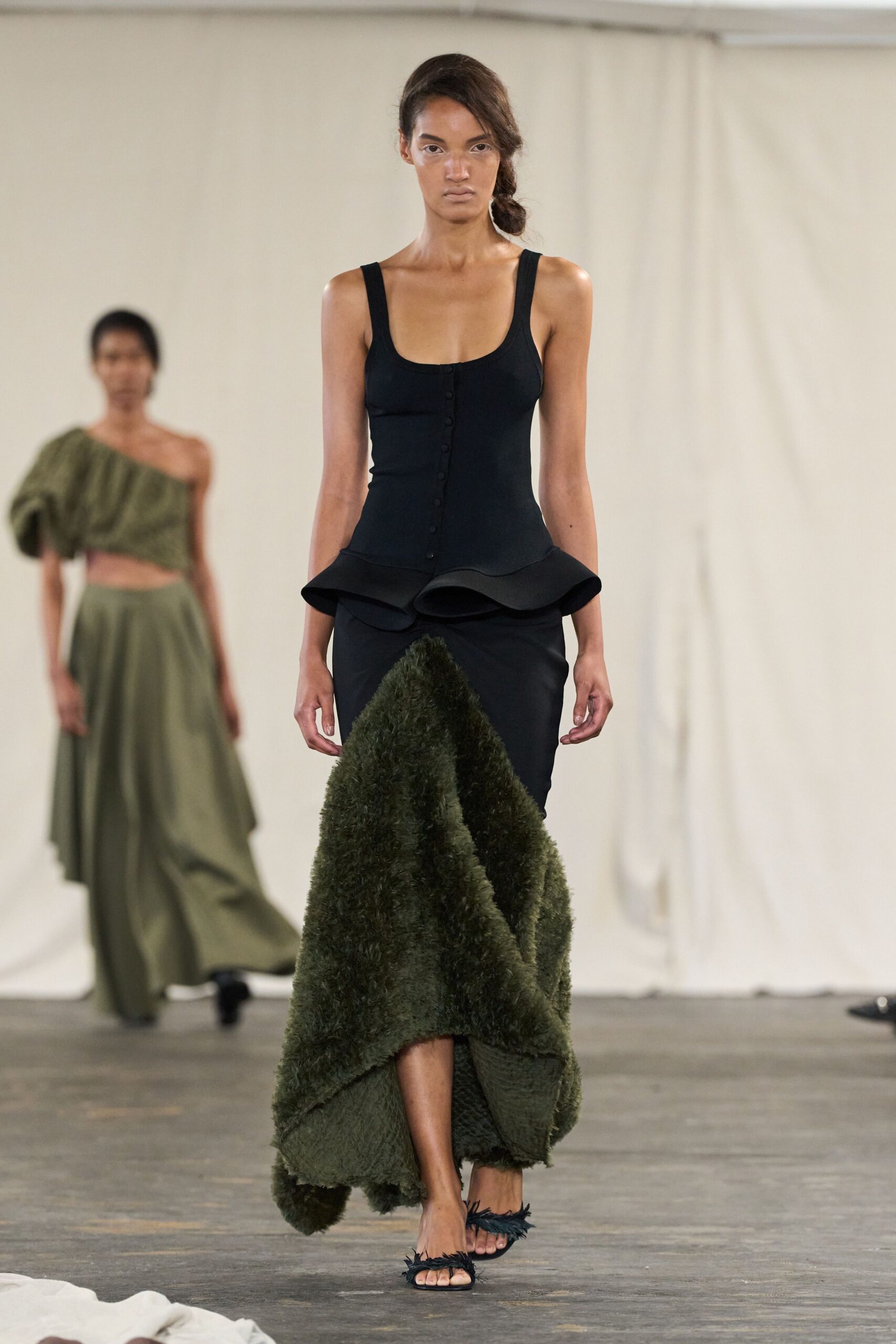 Black knit tank top, buttoned through the front with a flared peplum edge. Column skirt breaking into shaggy moss-green pile, longer and heavier in front. Black toe-ring sandals with feathery fringe across the straps. Walking out of a garden party after the grass has soaked through.