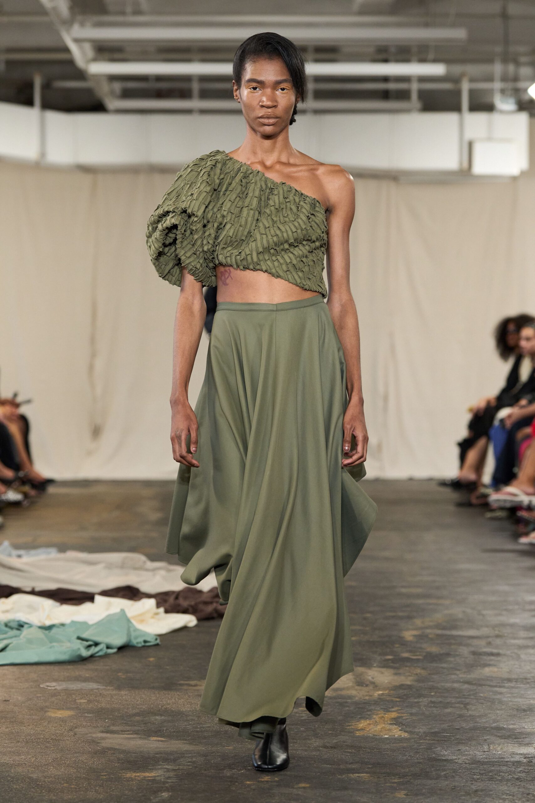Olive one-shoulder crop top in puckered smocking, one sleeve blown into a cocoon. Matching olive maxi skirt in fluid jersey, cut to fall from a plain waistband. Black leather ankle boots, squared at the toe. Walking through a conservatory after rain, hem skimming the damp stone path.