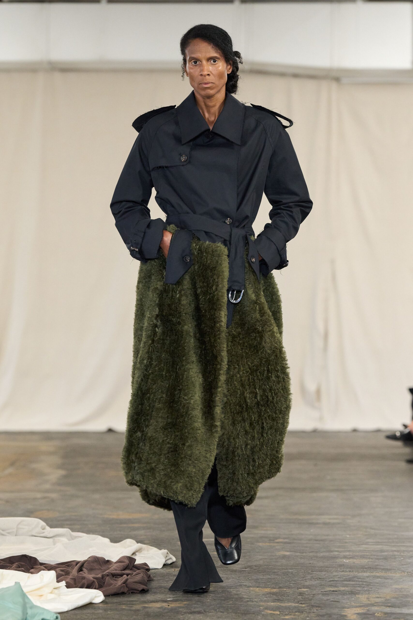 Black cotton gabardine trench top, cropped into an asymmetrical wrap with raw-fringed epaulettes. Deep moss shag-pile skirt swelling from the waist, almost blanket-thick. Black trousers breaking into slight flares over the instep. Square black leather toes. For crossing the yard at a drafty country house before dinner is called.