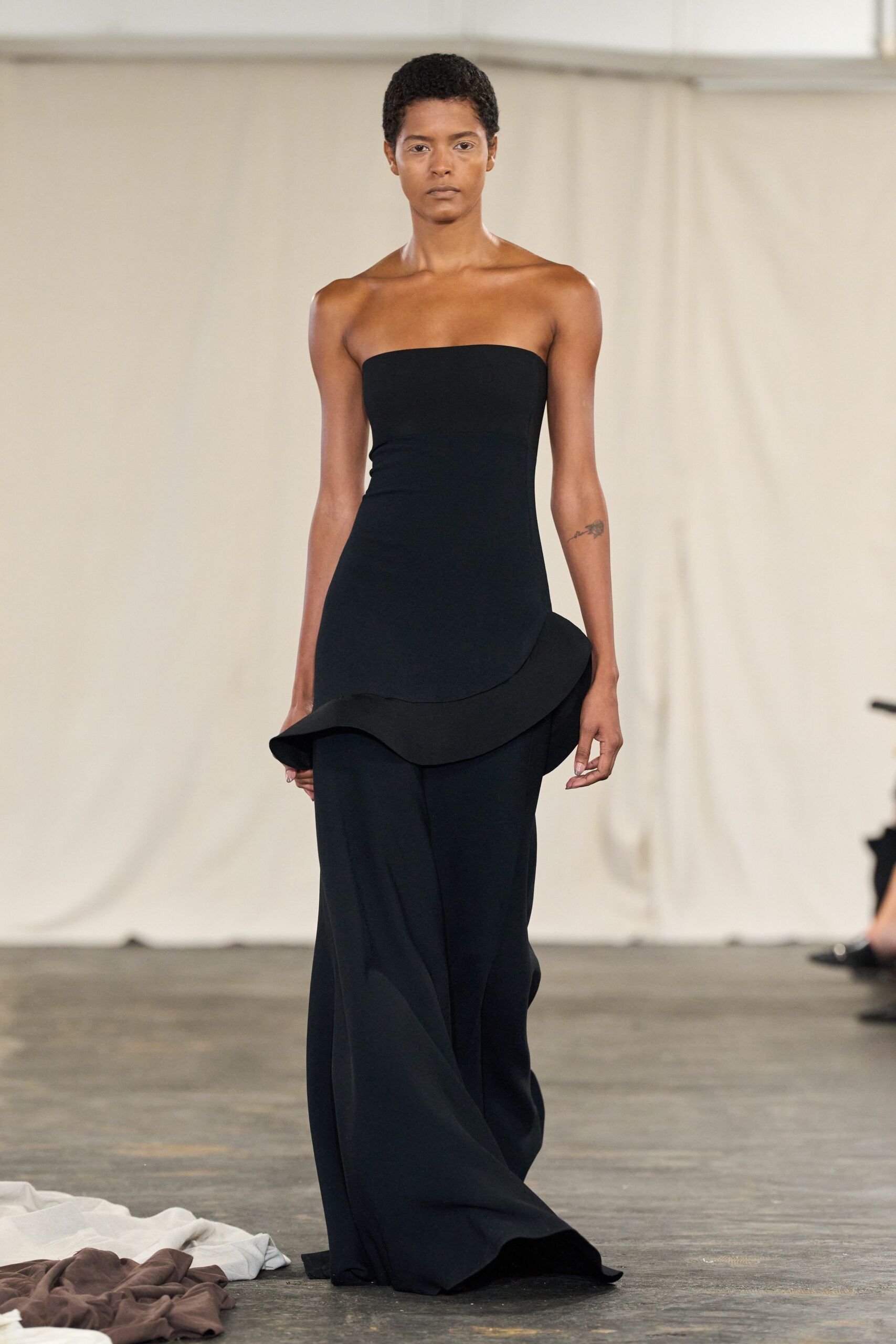 Black crepe column bodice with a wide sculpted flounce tipping at the hip. Floor-skimming black trousers in fluid cloth, breaking into a soft pooled hem. For crossing a bare hall after the speeches, heels clicking on concrete.
