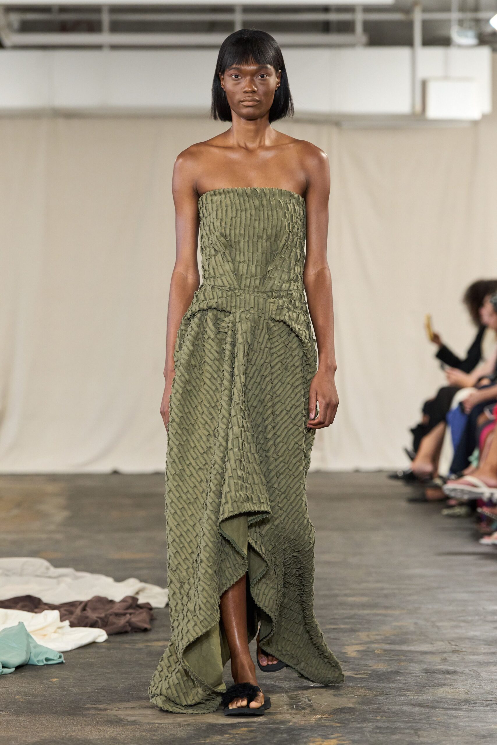 Strapless moss dress in puckered strips, draped into a raw-edged waterfall hem. Black flat sandals with fuzzy thong straps. Walking across a concrete studio floor after the dye vats have cooled.