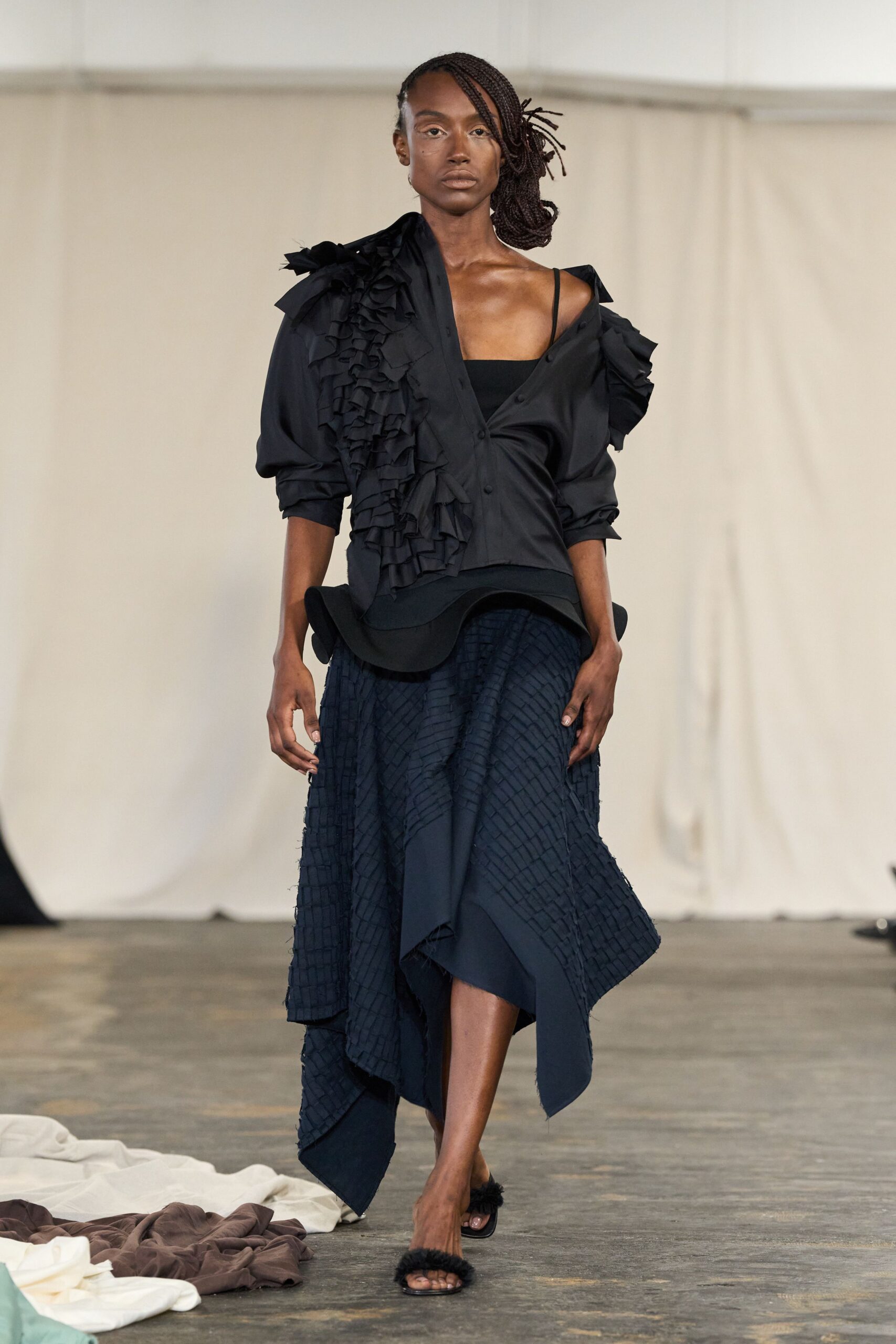 Black blouse with dense ruffles cascading from shoulder to placket. Straight black camisole, one thin strap exposed off the shoulder. Asymmetric midi skirt in frayed grid-textured cloth, hem dropping to a point. Black slide sandals with fuzzy straps. Walking across a concrete hall after rain, still carrying the night's heat.