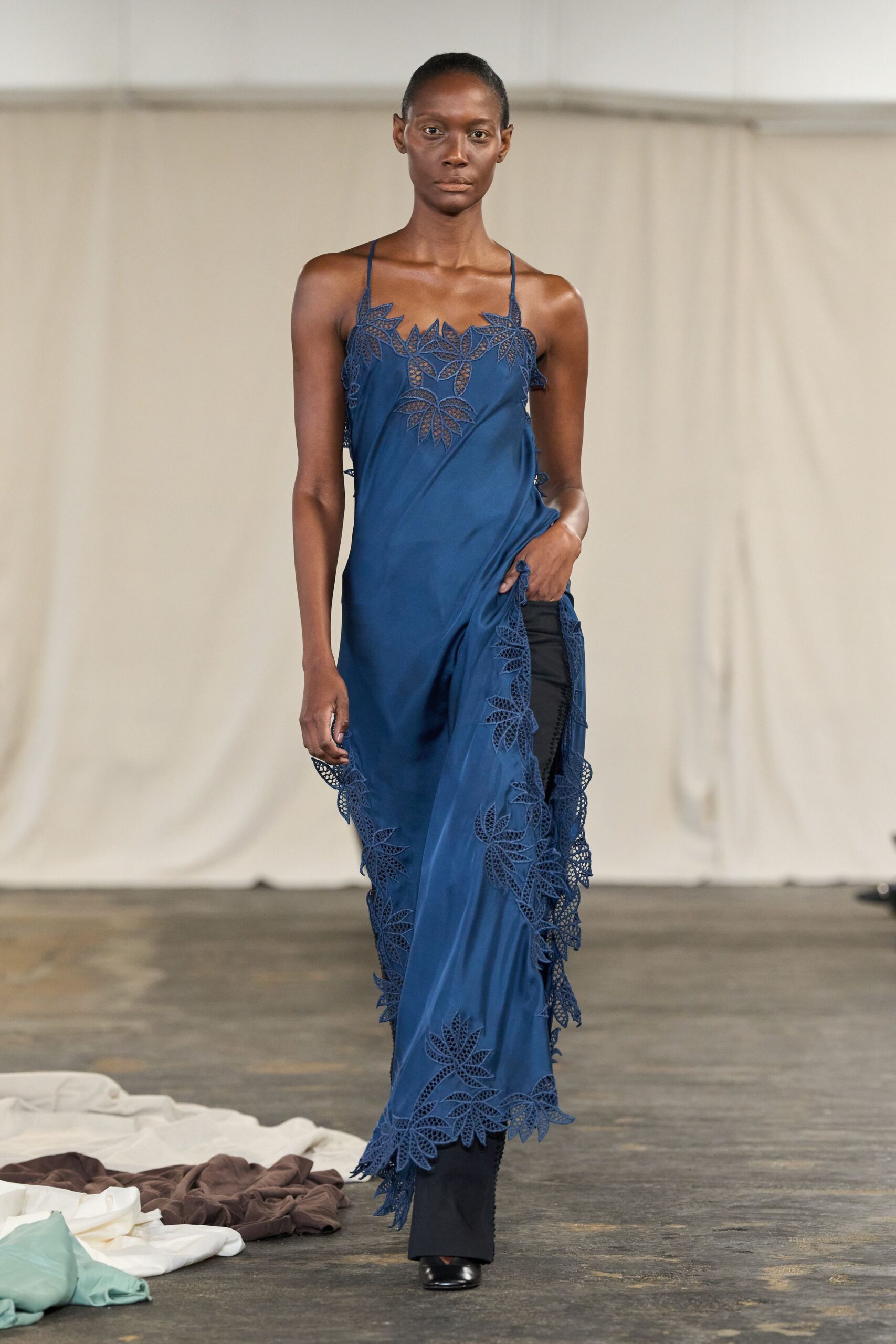 Ink-blue slip dress with leaf-cut lace climbing the neckline and hem. Black straight trousers showing through the thigh-high slit. Black square-toe shoes, only the vamp flashing forward. Walking across a hotel courtyard after rain, hem skimming wet flagstones.