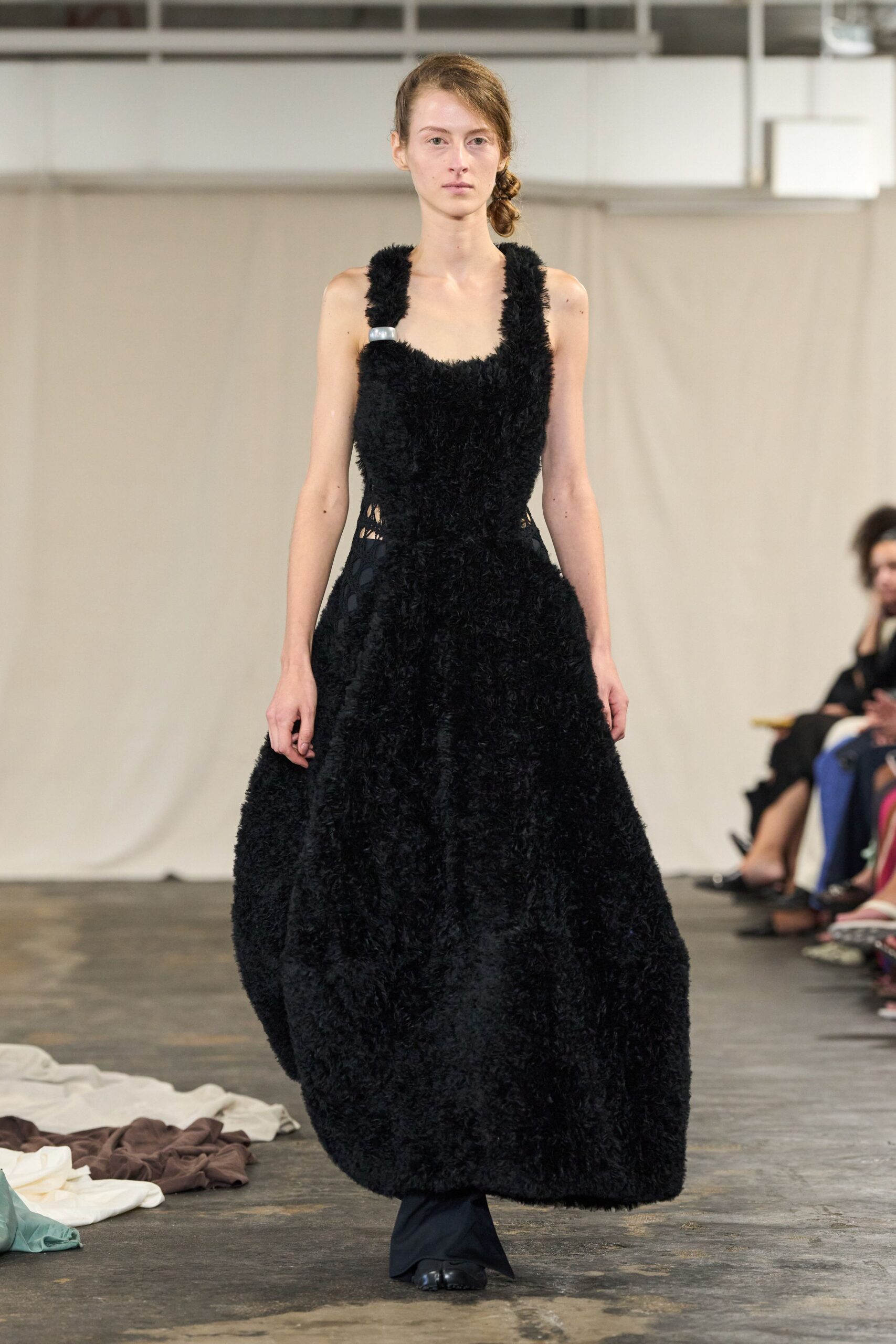 Black shag-pile dress with a scooped bib front and swollen skirt. Black trousers peeking beneath the hem, pooling lightly over the shoe. Square black flats with a split toe. For crossing a concrete studio floor after everyone else has gone home.