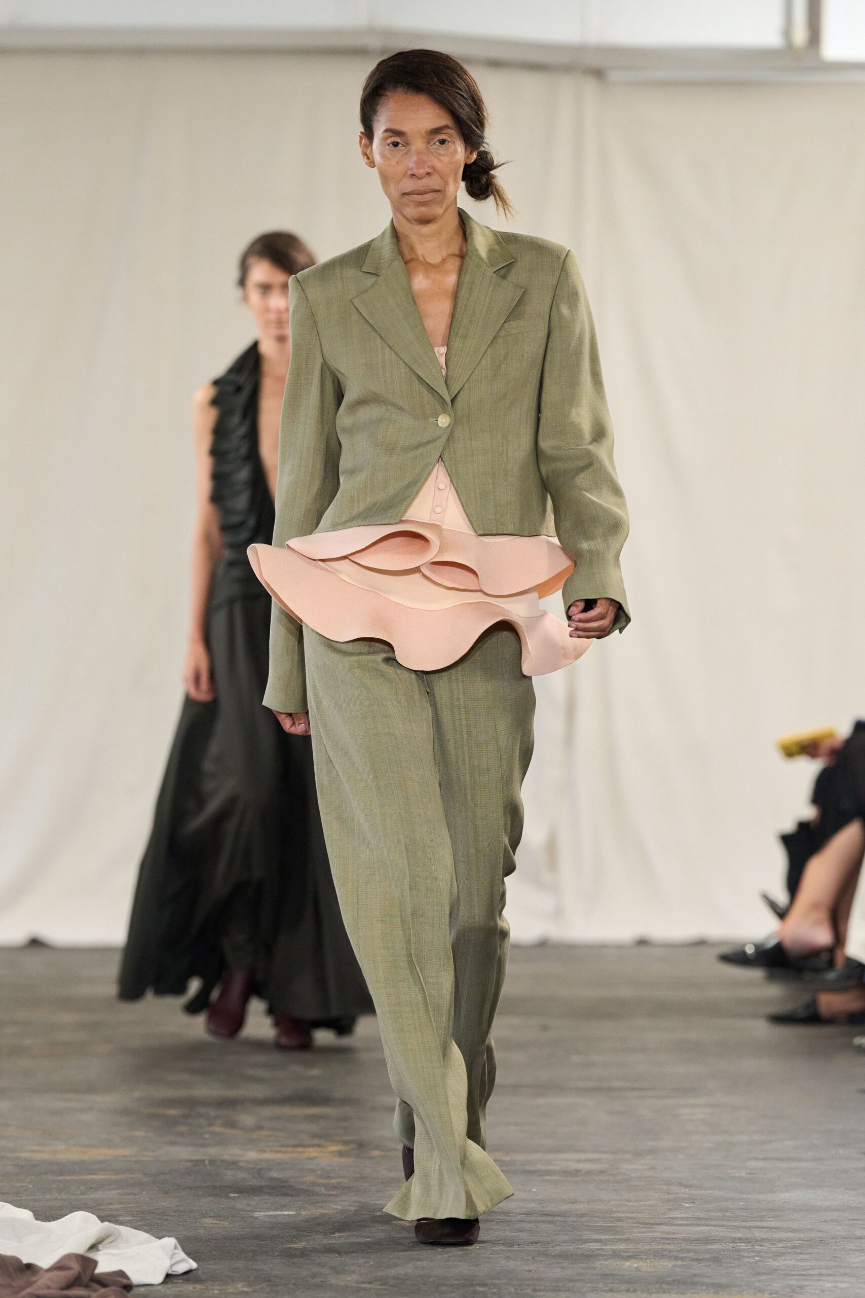 Sage suiting blazer, cut short with one low pearl button. Blush top exploding into thick sculpted ruffles at the hips. Matching straight trousers in the same dry slub weave. Dark pointed boots peeking under the hem. For signing papers in a drafty municipal office with flowers already collapsing.