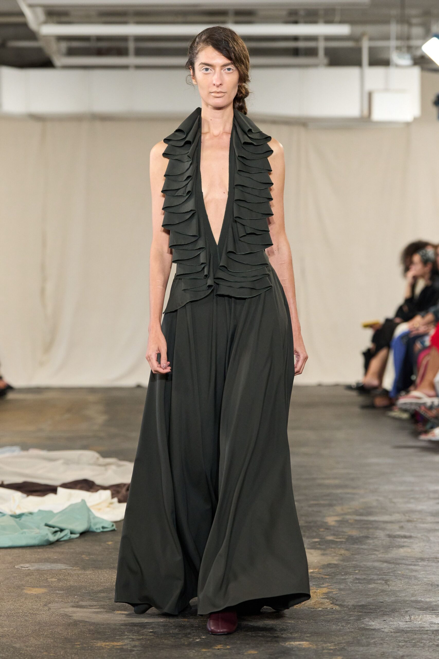 Black crepe jumpsuit with a plunging halter front and stacked ruffle lapels. Palazzo legs falling from a gathered waist, hems nearly grazing the floor. Square-toe oxblood boots peeking out beneath the sweep. Crossing a concrete loft with fabric bolts kicked into the aisle.