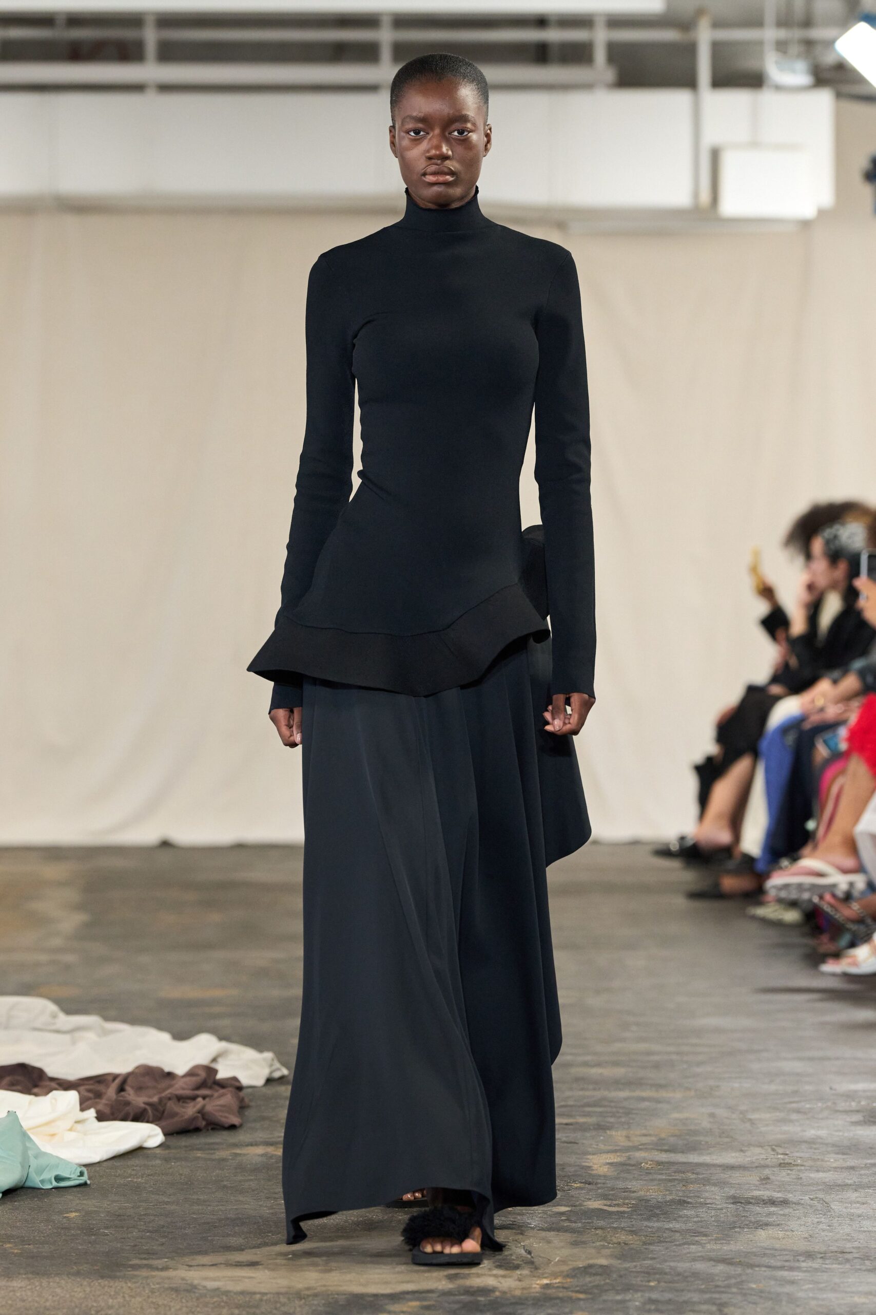 Black knit turtleneck, body-skimming with a flared peplum cut on the slant. Inky fluid maxi skirt falling in broad panels to a dragging hem. Flat black sandals with a fuzzy vamp band. For crossing a concrete hall before sunrise, carrying your shoes home.