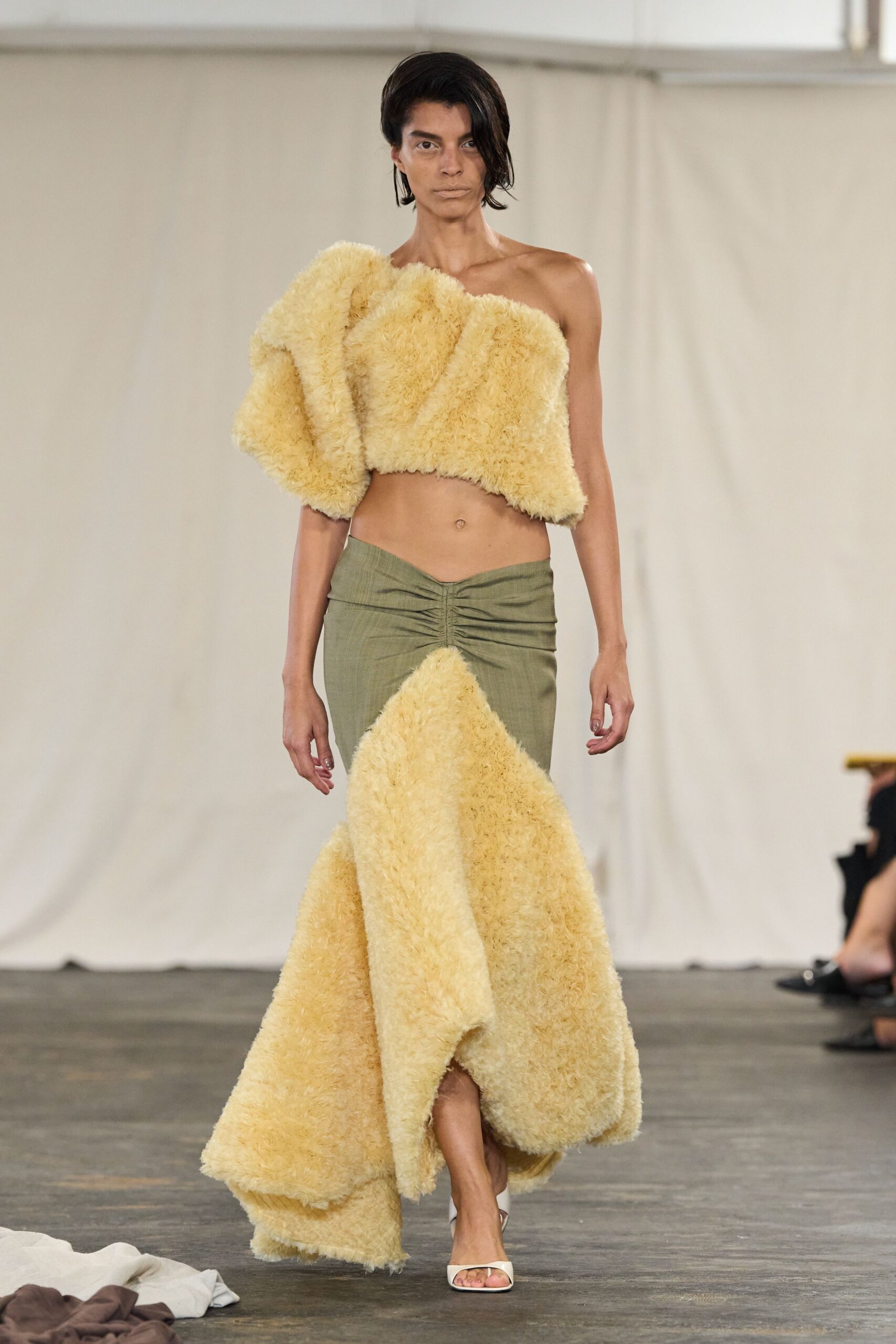 Buttercream shaggy one-shoulder crop top, folded into oversized padded swells. Olive low-slung skirt, ruched at center with a shaggy triangular godet. White square-toe mules. For crossing a hotel lobby still damp from the pool.