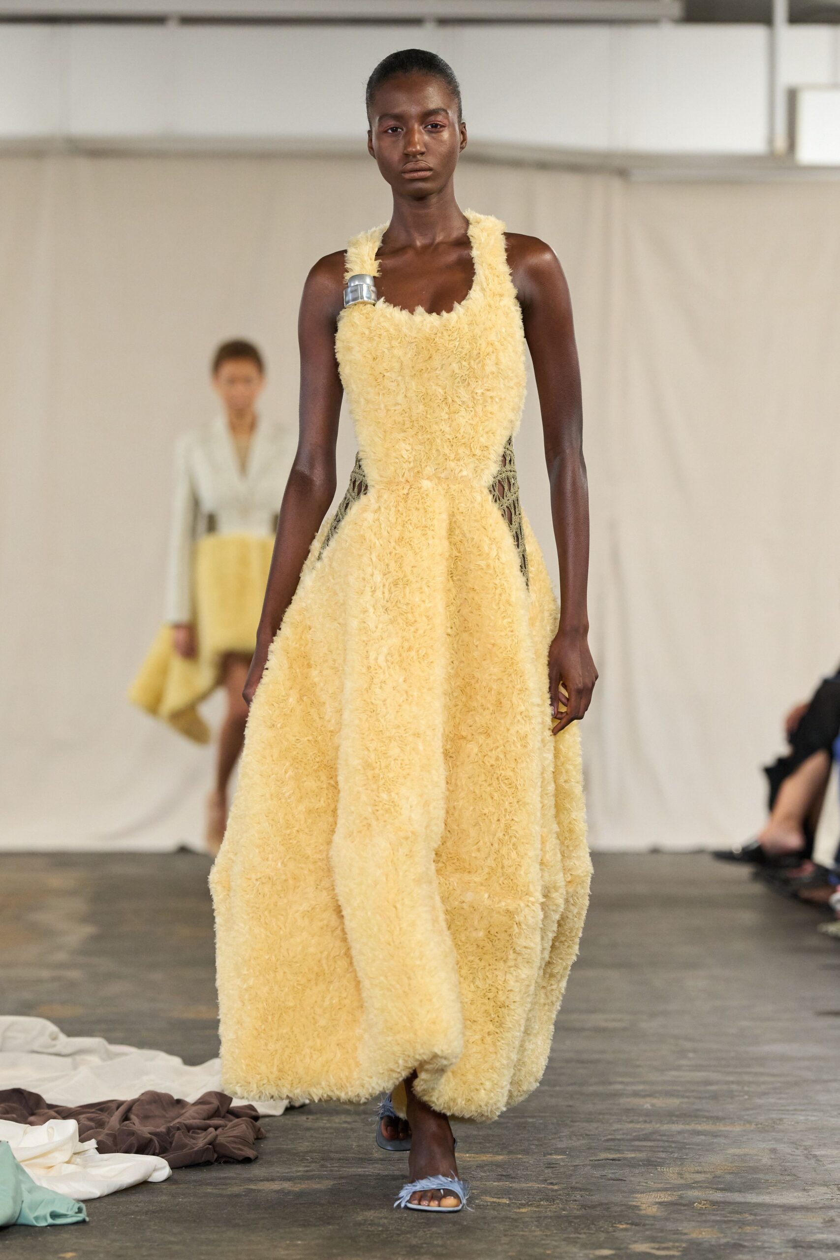Butter-yellow shaggy gown with a scooped neck and black openwork side panels. Single brushed-silver cuff clamping the left shoulder strap. Pale blue feather-trim slides peeking from the swollen hem. Crossing a concrete hall after someone spilled armfuls of fabric samples.