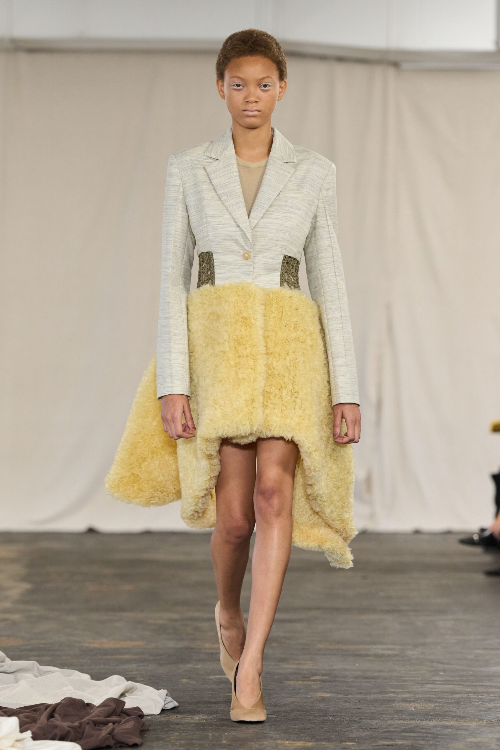 Pale grey tailored bodice spliced into a butter shearling skirt, one horn button. Sheer beige mesh at the neckline. Nude suede pumps with narrow pointed toes. Walking across cold studio concrete while the space heaters are still losing.