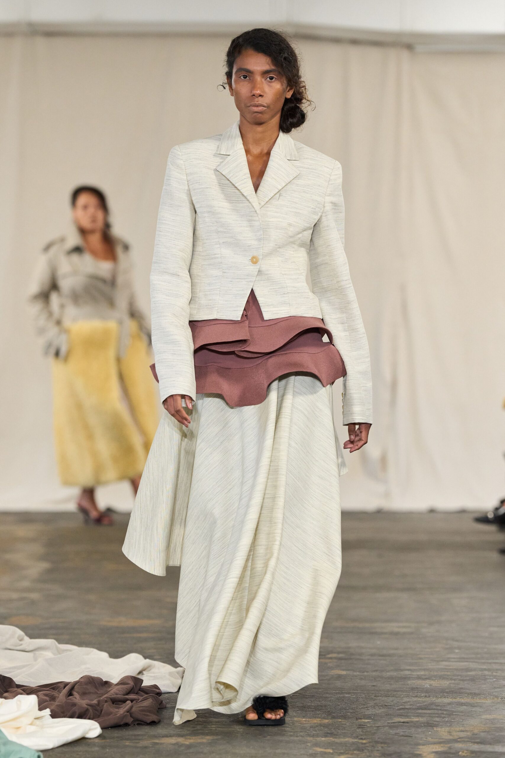 Pale slub-weave jacket cut close, fastened with one honey button. Dusty mauve peplum flaring in stacked scalloped layers. Matching cream slub skirt draped into a long off-centre fall. Black furry slide sandals. Crossing a drafty hall with the tablecloths still pooled on the floor.