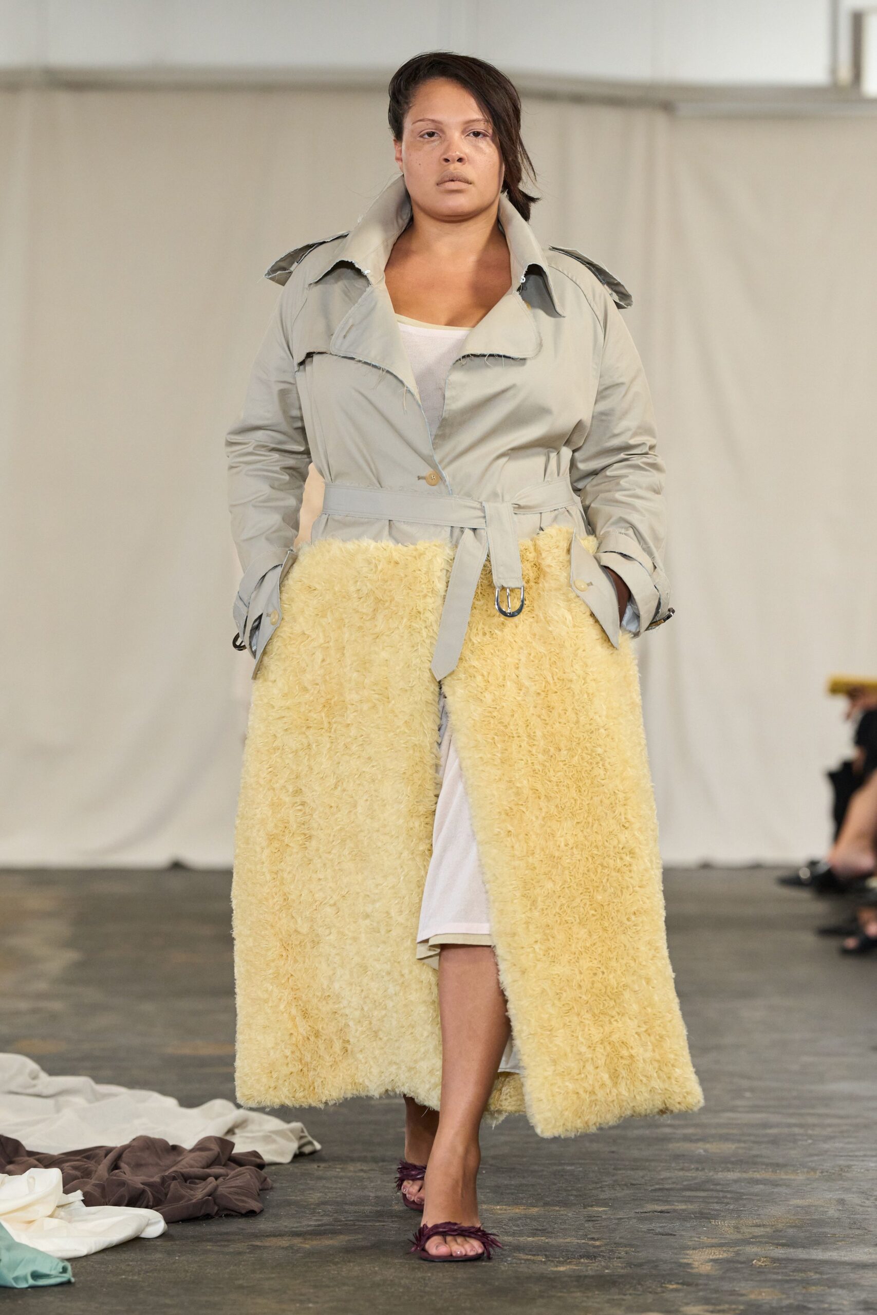 Pale grey trench spliced to a butter shag lower half, collar edges left raw. White slip peeking through the front split. Plum feather-trim thong sandals. Walking across a drafty studio floor while the dropped cloths stay underfoot.