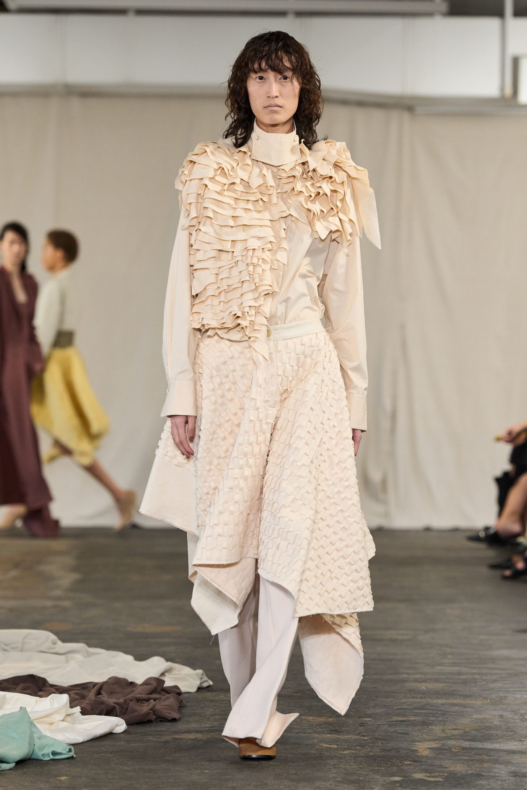 High-neck cream blouse smothered in cascading ruffles across one shoulder. Cream wrap skirt in small woven tabs, hem dropping in uneven points. Pale fluid trousers falling straight beneath the skirt. Square-toe pumps in tan and black. Walking through a silent textile studio before the steam irons start.