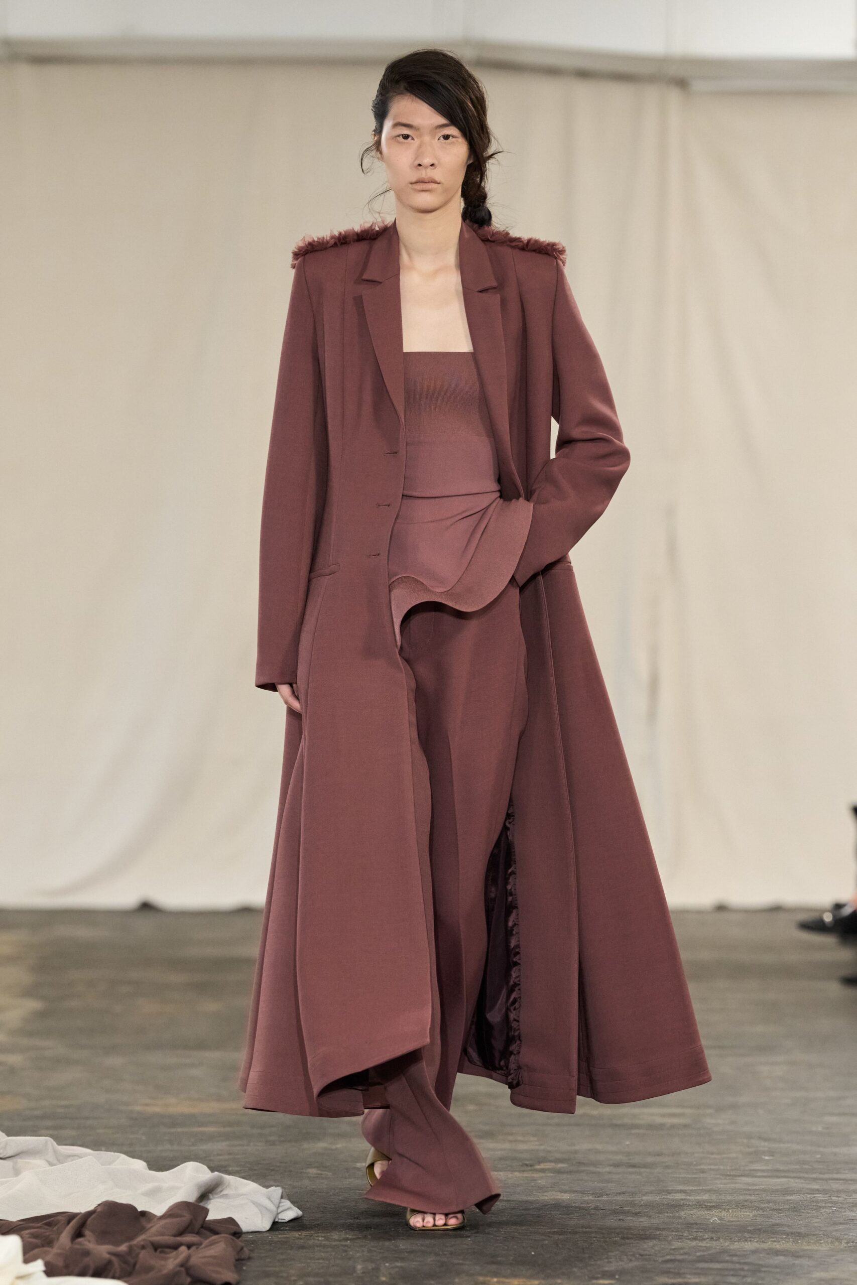 Long oxblood coat in dense crepe, shoulders tufted with short fringe. Matching bandeau top with a clean squared edge. Fluid wide-leg trousers with a folded drape spilling over the hip. Barely there heeled sandals. For crossing an echoing hall after the speeches have finished.