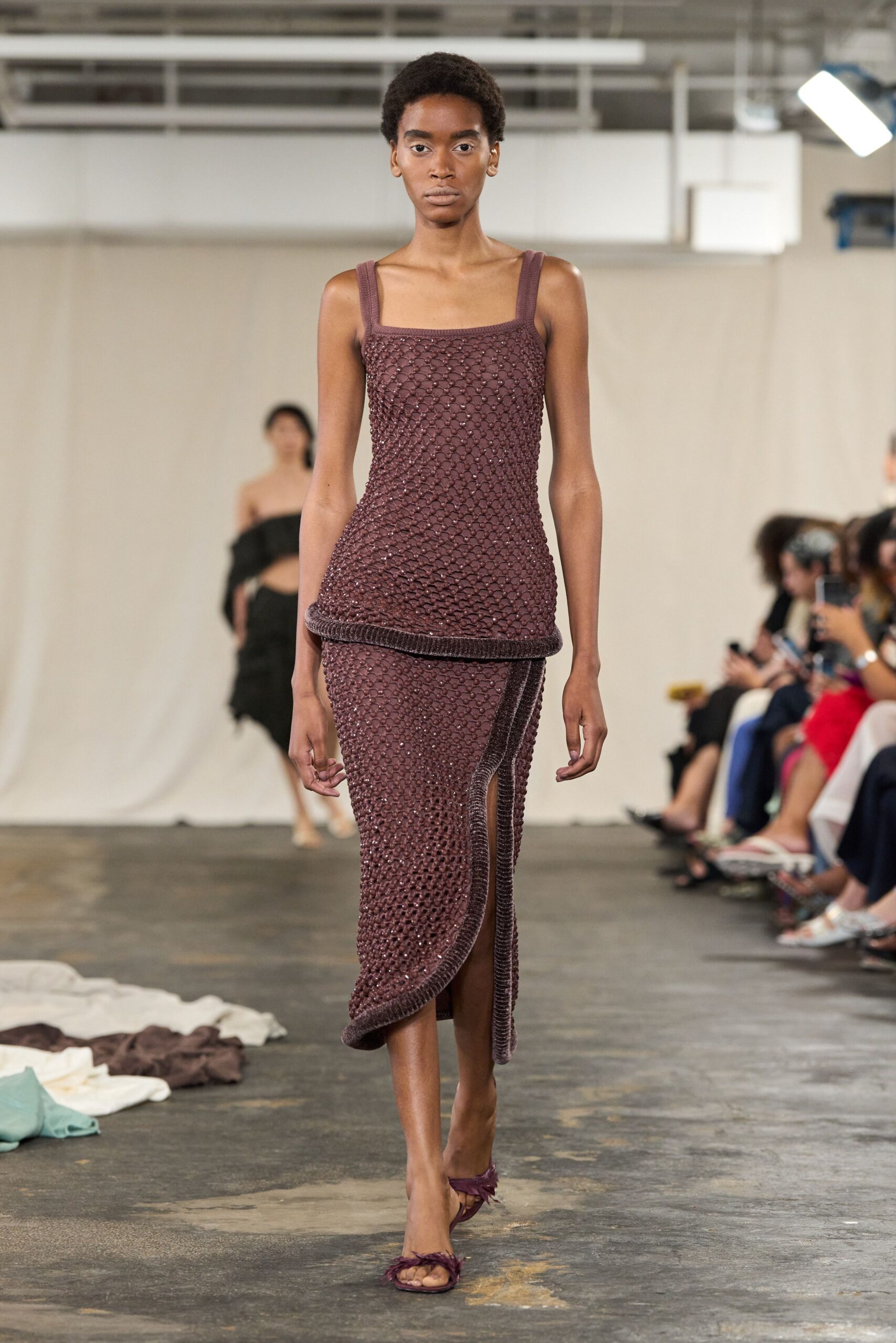 Mulberry openwork knit tank dress, looped surface over a fitted underlayer. Matching wrap panel edged in thick ribbing, one side hanging lower. Plum thong sandals with feathery tufts across the toes. Walking across a cooling terrace after the wedding plates are cleared.