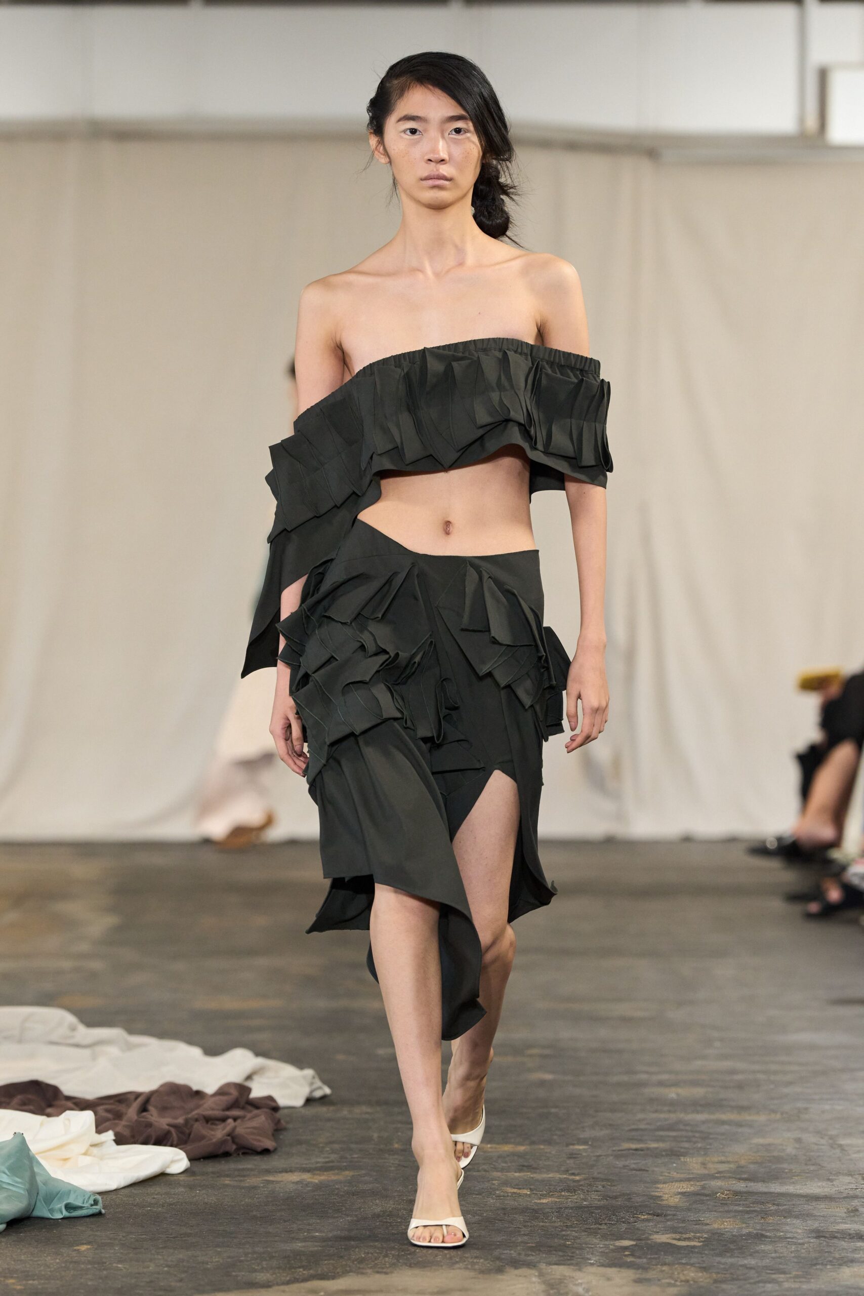 Black taffeta bandeau with accordion flounces slipping off one shoulder. Asymmetric black skirt, layered with folded taffeta shards and a thigh slit. White square-toe mules. Walking across a half-cleared studio floor after everyone else has gone.