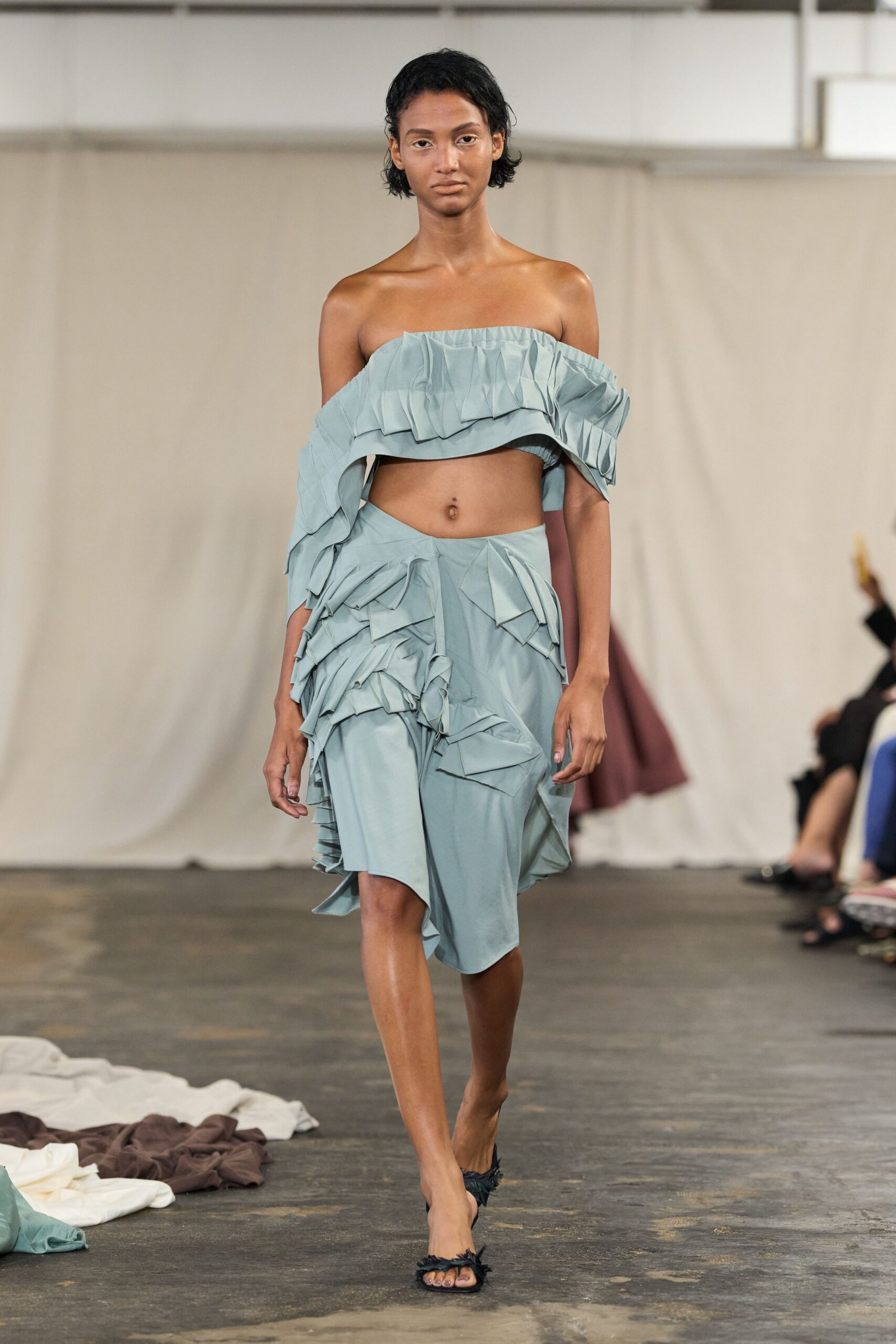 Pale celadon bandeau top with crisp folded ruffles slipping off the shoulders. Matching wrap skirt, tiers of origami-like flounces bunching at one hip. Black slide sandals with frayed petal trims across the toes. Walking across a concrete gallery after someone shook out the table linens.