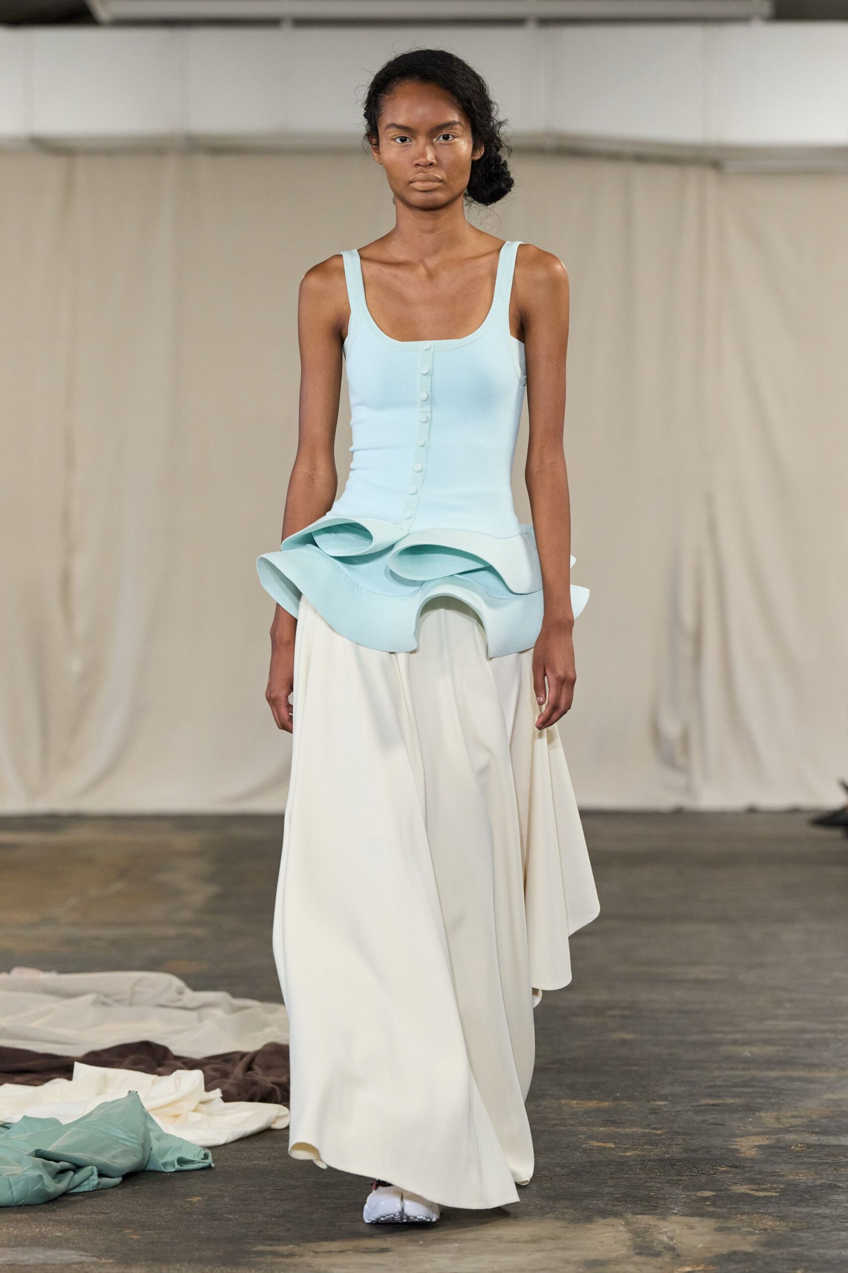 Powder-blue stretch top with covered buttons and a rigid looped peplum. Milk-white skirt falling in heavy fluid folds to the floor. White split-toe flats peeking under the hem. Crossing a museum atrium while the movers are still uncrating sculpture.