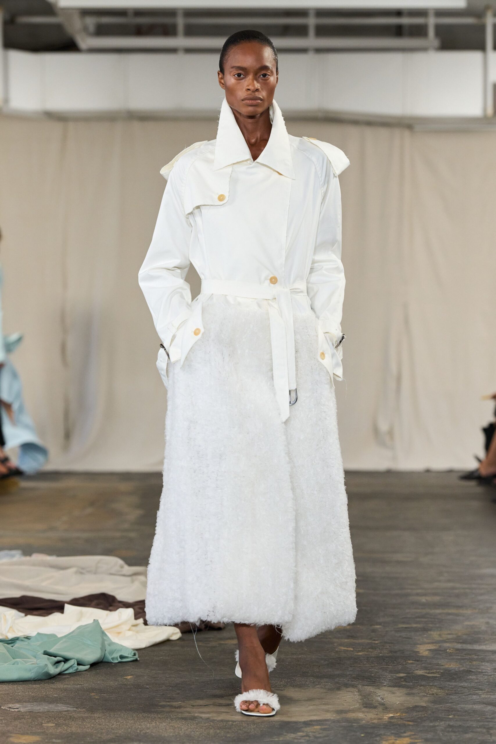 Ivory trench coat with a high stand collar and offset horn-look buttons. Long white boucle skirt, shaggy at the hem with a loose thread trailing. White open-toe mules banded in fuzzy trim. For crossing a concrete studio floor while the dye lots dry nearby.